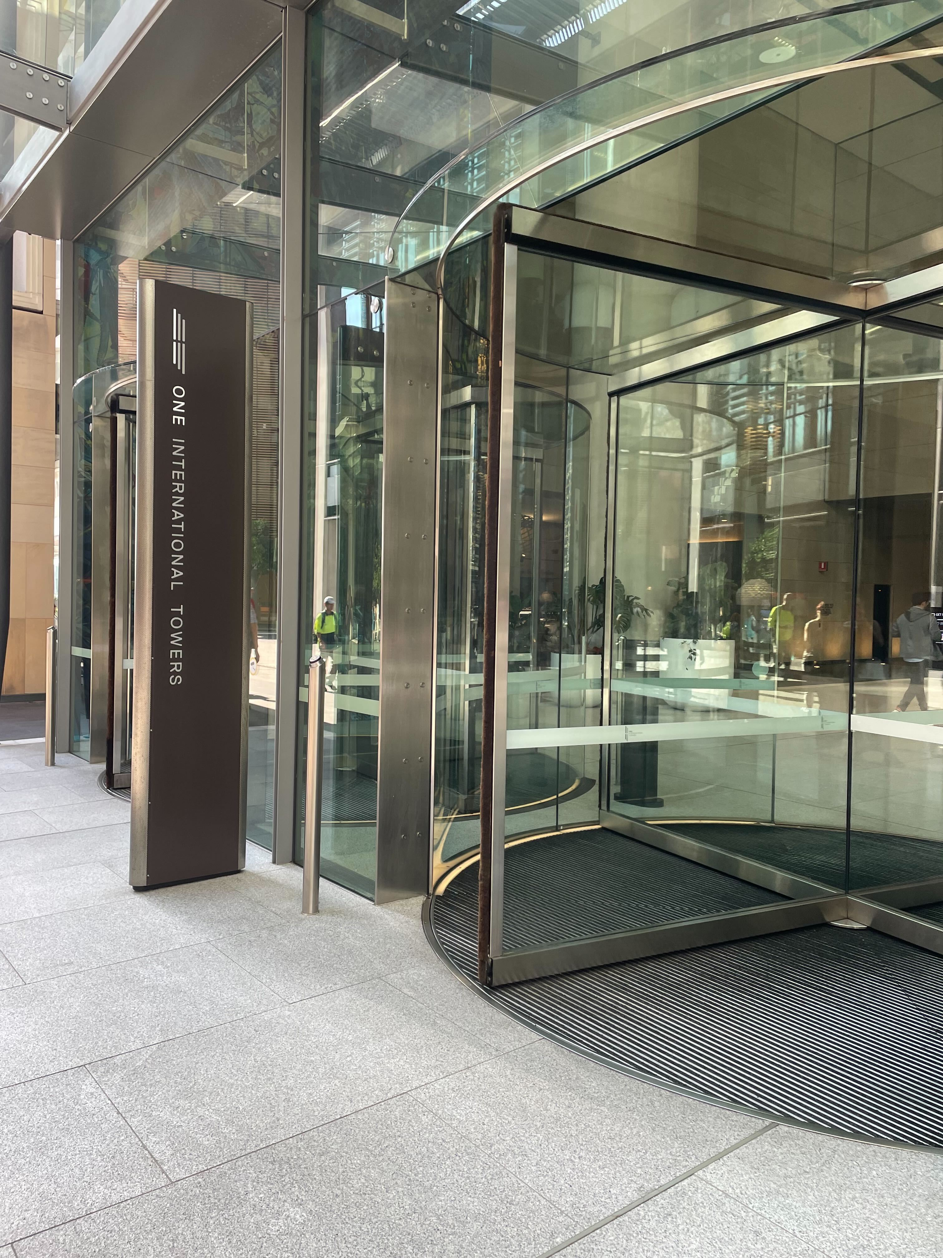 Image of glass revolving doors with a grey sign next to it which reads 'One International Towers'. 