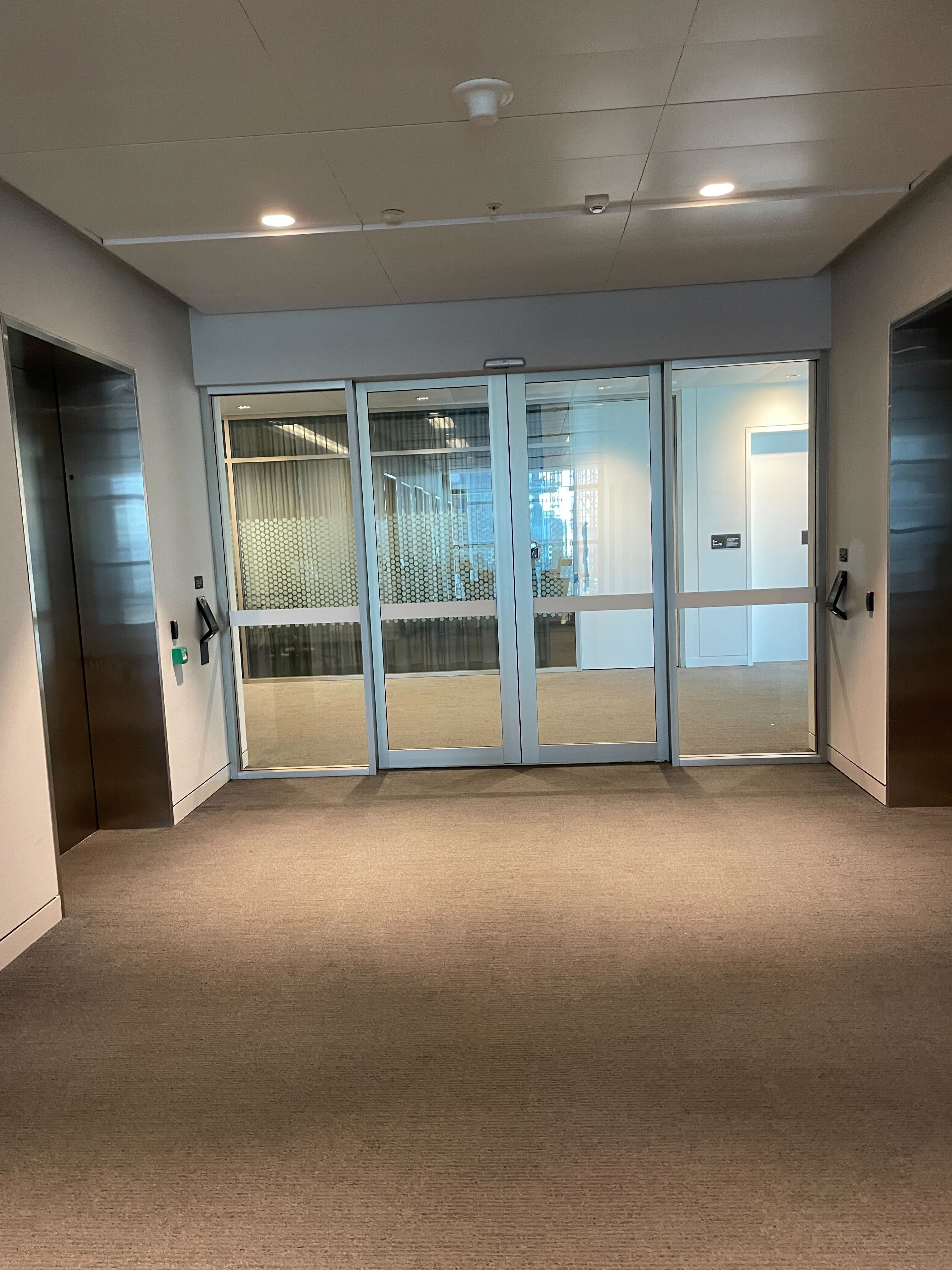 Image of corridor of lifts and sliding glass doors that open with a key card to another corridor. The floor is grey and the walls are white 