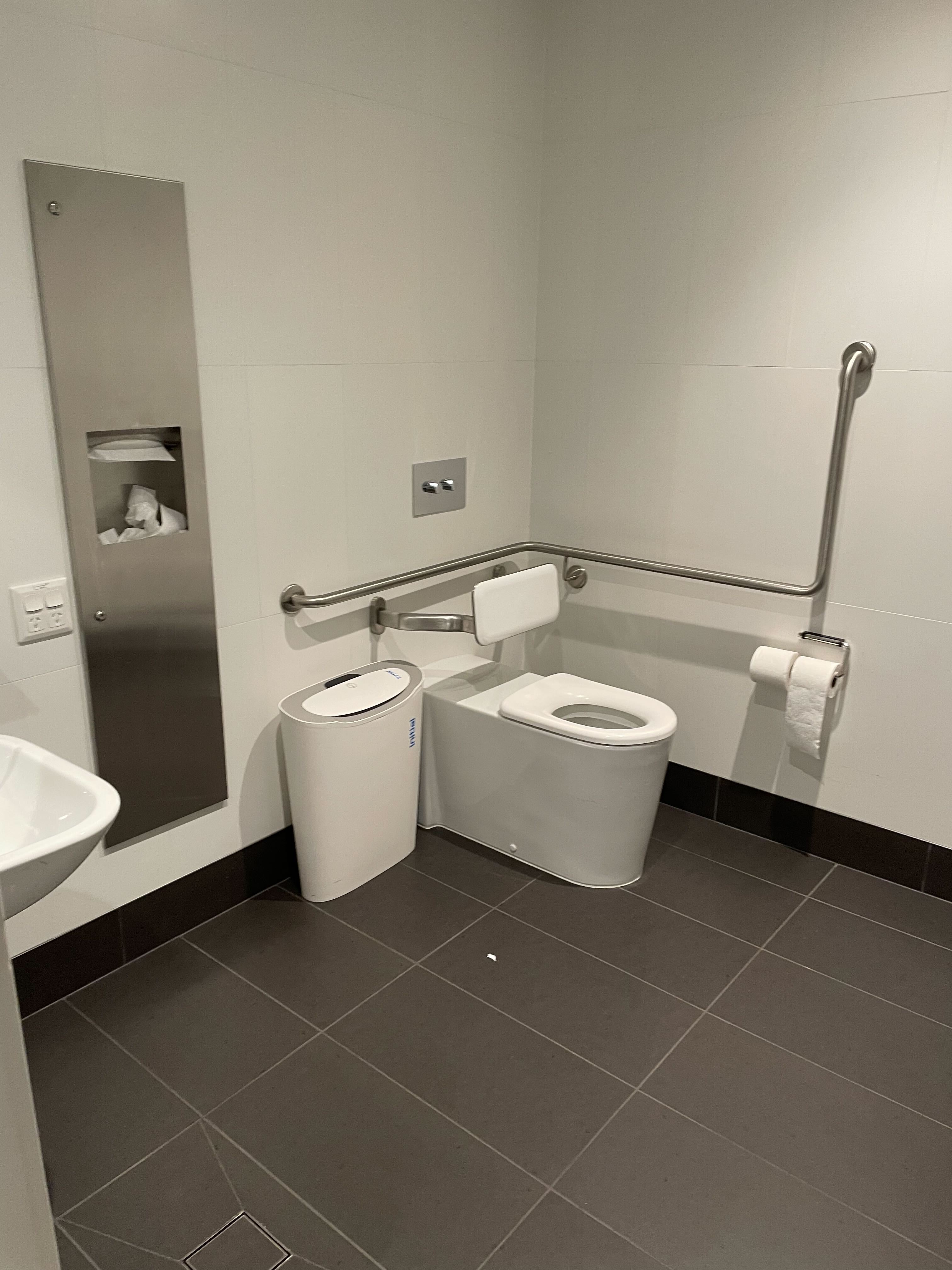 Image of inside the bathroom. The floor tiles are black, and the wall tiles are white. The toilet has railings attached, a sanitary bin and a sink with wheel space below. 