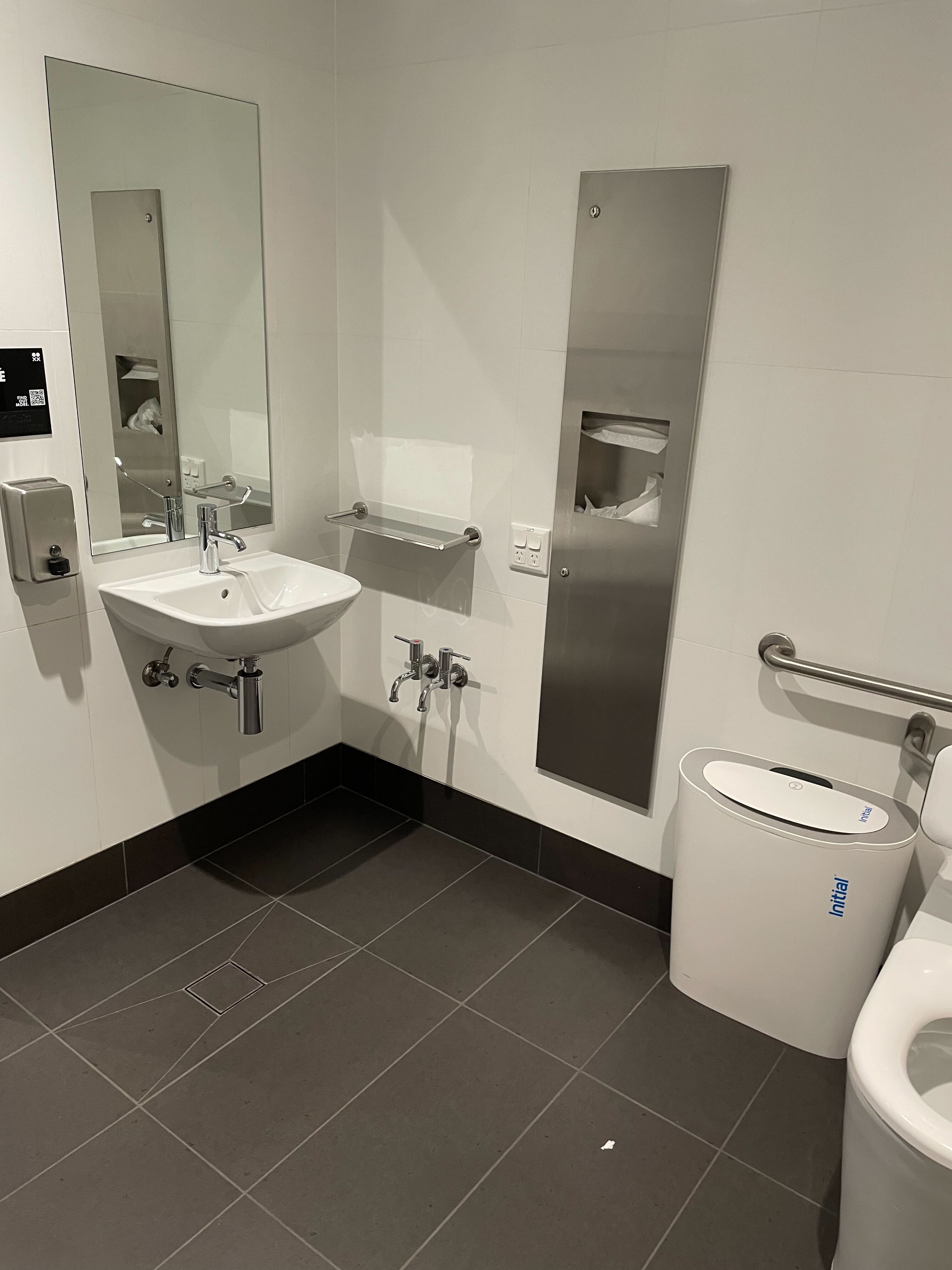 Image of the other side of the bathroom. The floor tiles are black, and the wall tiles are white. The toilet has railings attached, a sanitary bin, a sink with wheel space below, and paper towels. 
