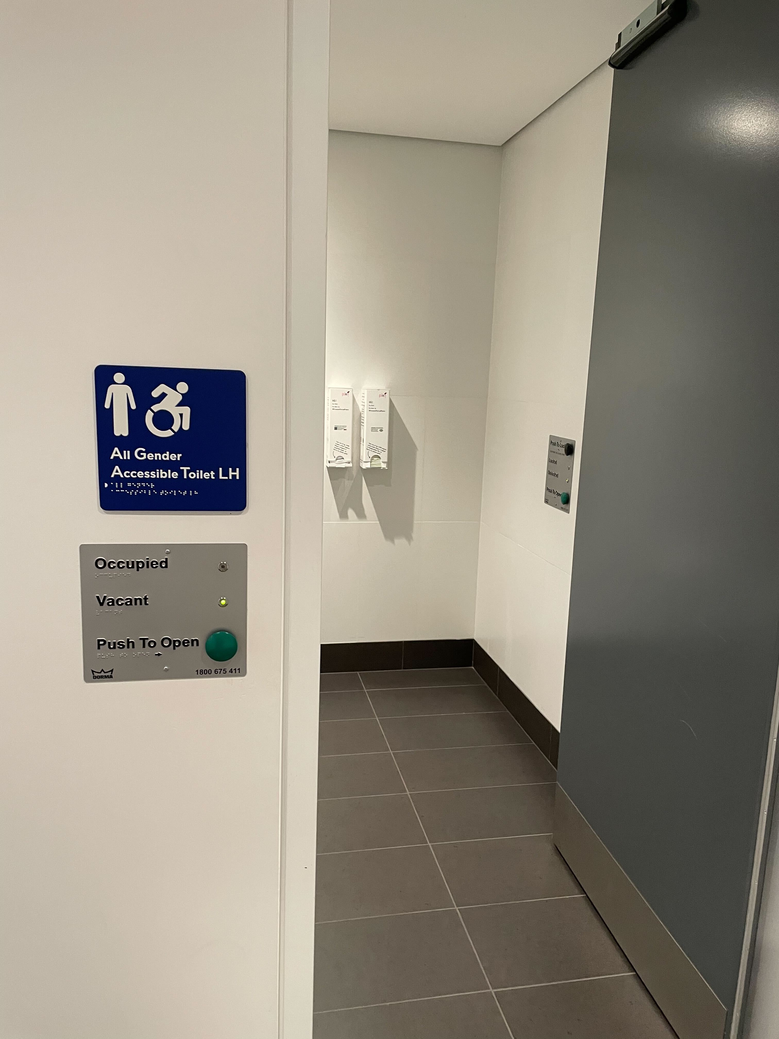 Image of accessible staff bathroom. There is a wide black door, black floor tiles, and white wall tiles. A blue sign reads 'All Gender Accessible Toilet LH'. Below is a button to open the door, then make the bathroom occupied and vacant.