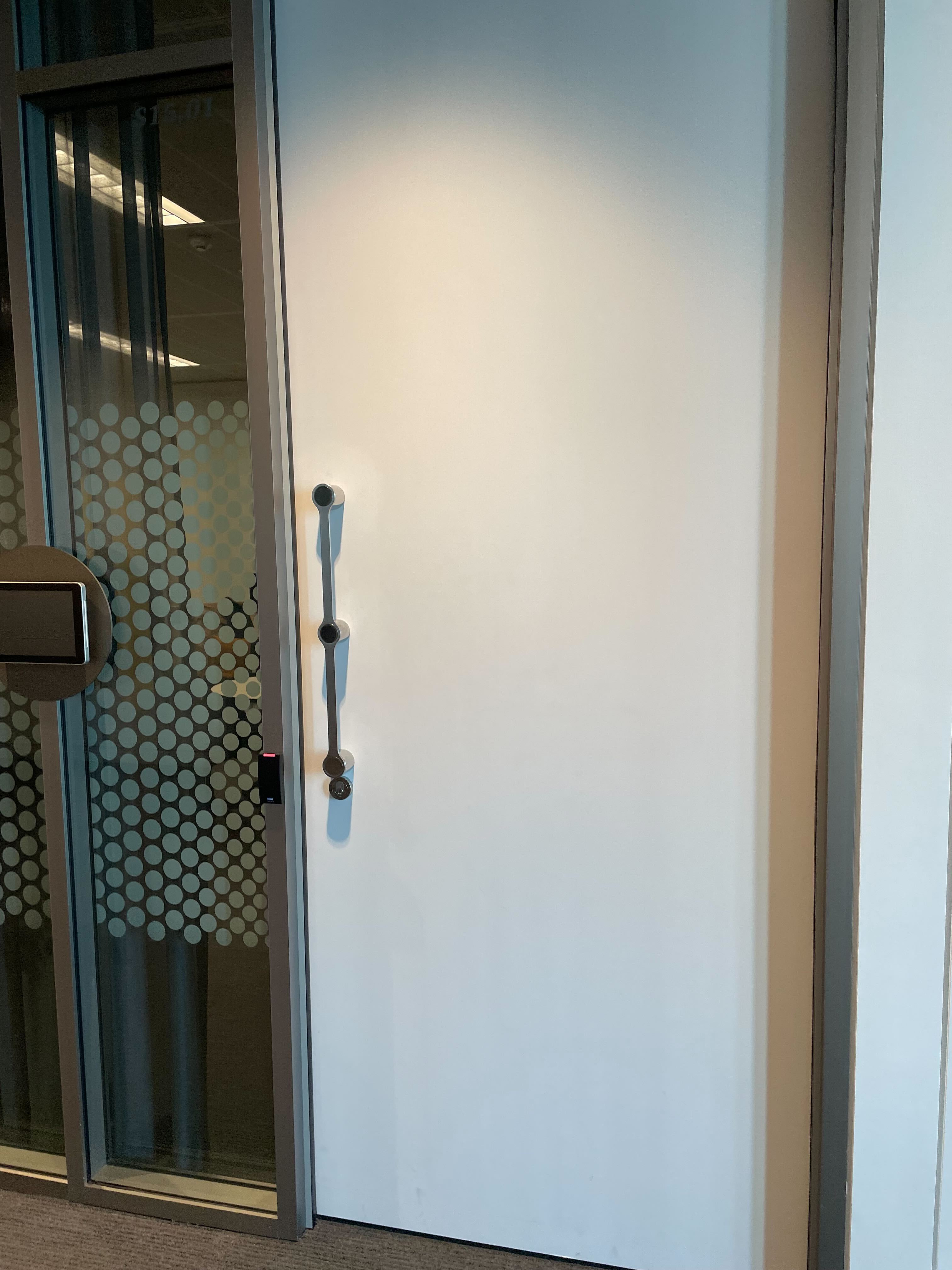 Image of the white office door that can be opened by a swipe card. The door has a long metal handle, with a spotted office window next to it. 