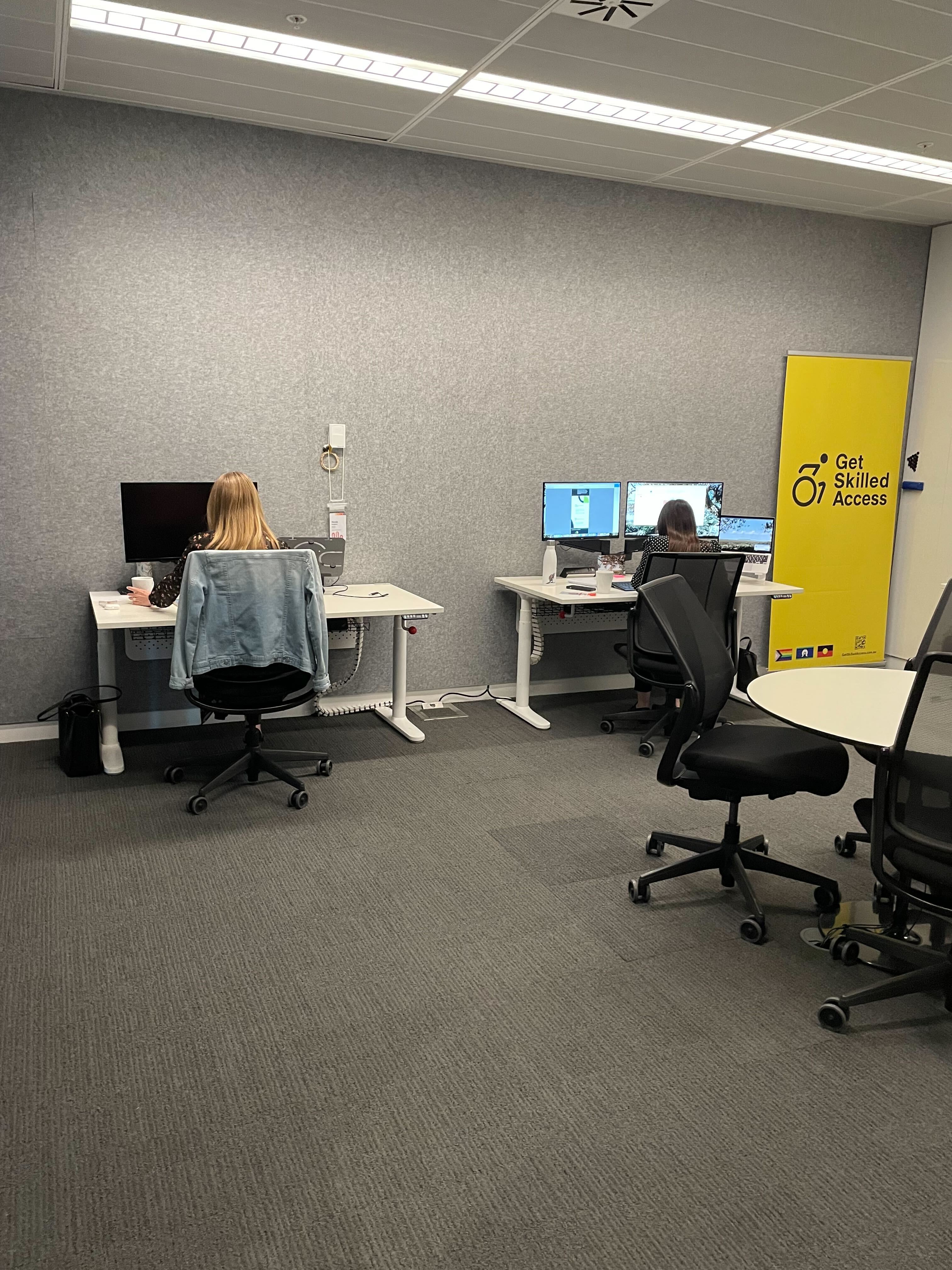 Image of two GSA staff members at their desks. The desks have laptops and screens. There is a spare desk in the center of the room. A yellow GSA sign sits in the corner of the room. 