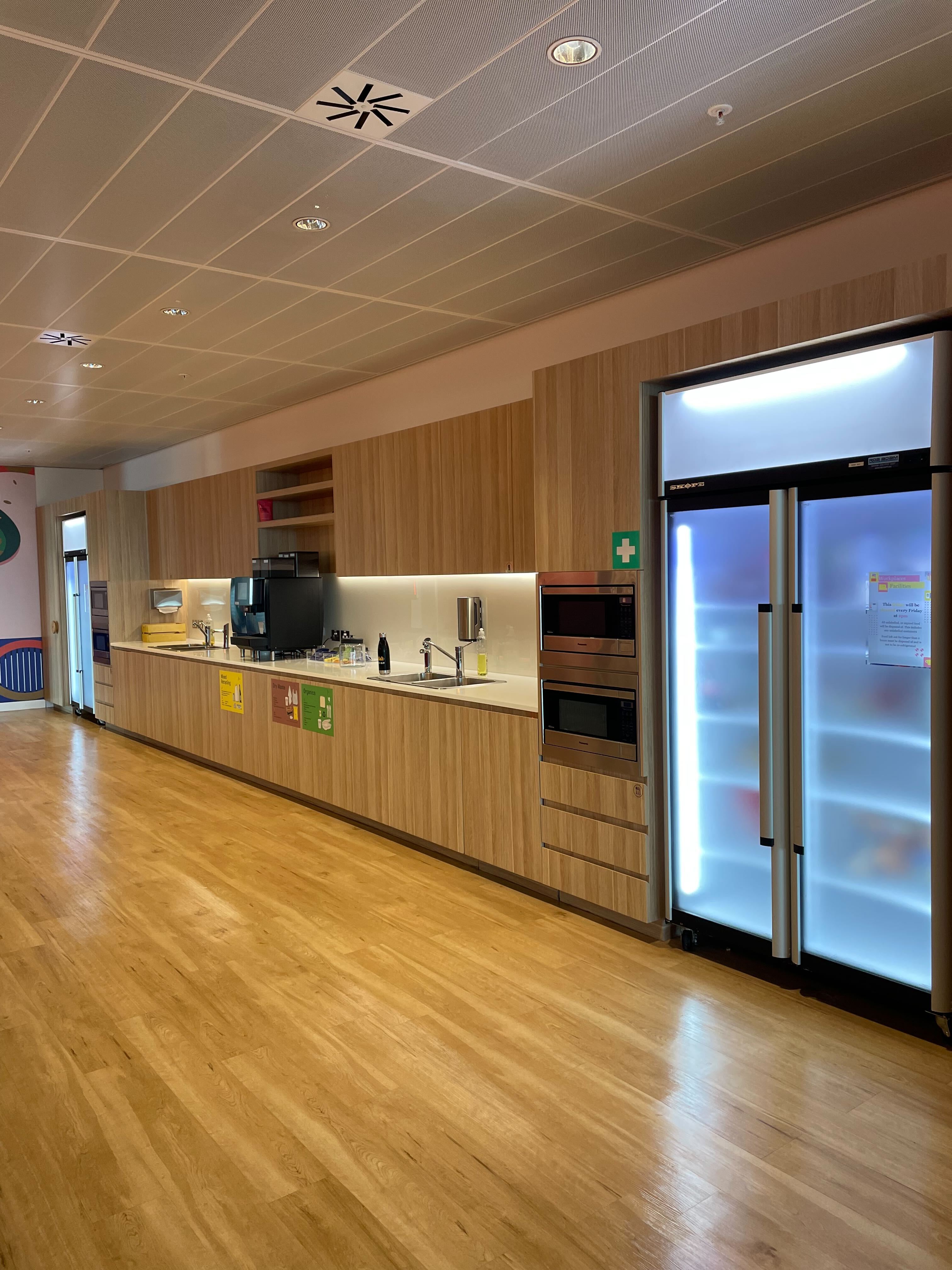 Image of a long wood paneled kitchen. There are two glass fridges, 4 microwaves, a coffee machine and two sinks. 