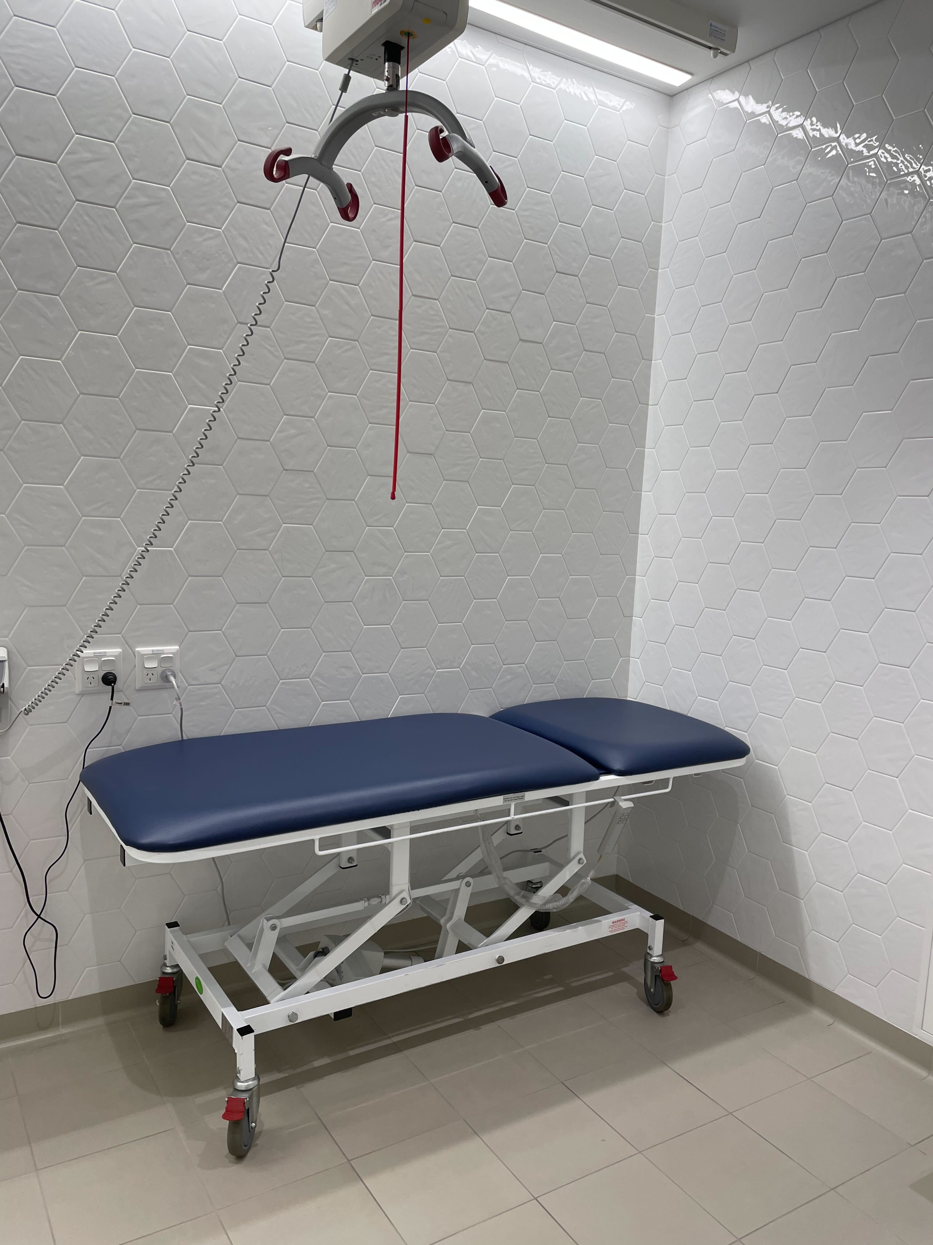 Image of inside the accessible change room. The floor tiles are grey, and the wall tiles are white. There is a blue change table with a hoist above.  