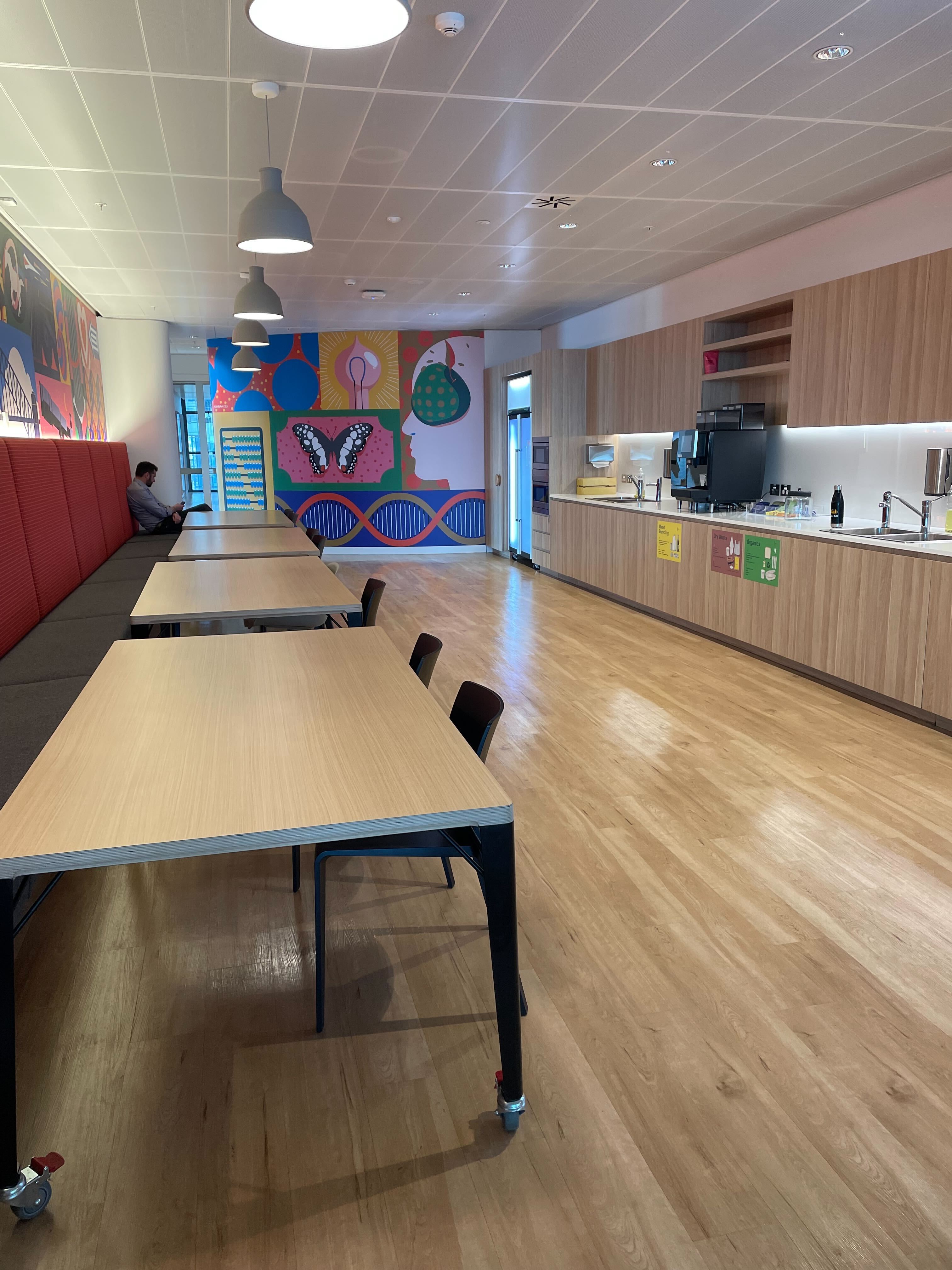 Image of the other side of the kitchen. There are four long wooden tables with black seats on one side of them and red and grey booth seating on the other.