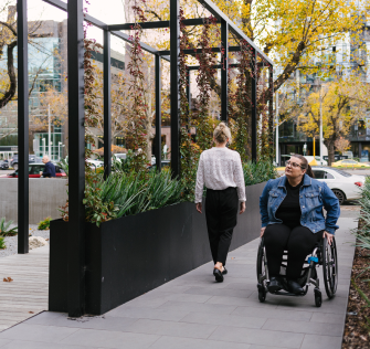 A Caucasian woman with dark hair, wearing glasses, is in a wheelchair, rolling towards a nearby building. Next to her, a woman is walking in the opposite direction.