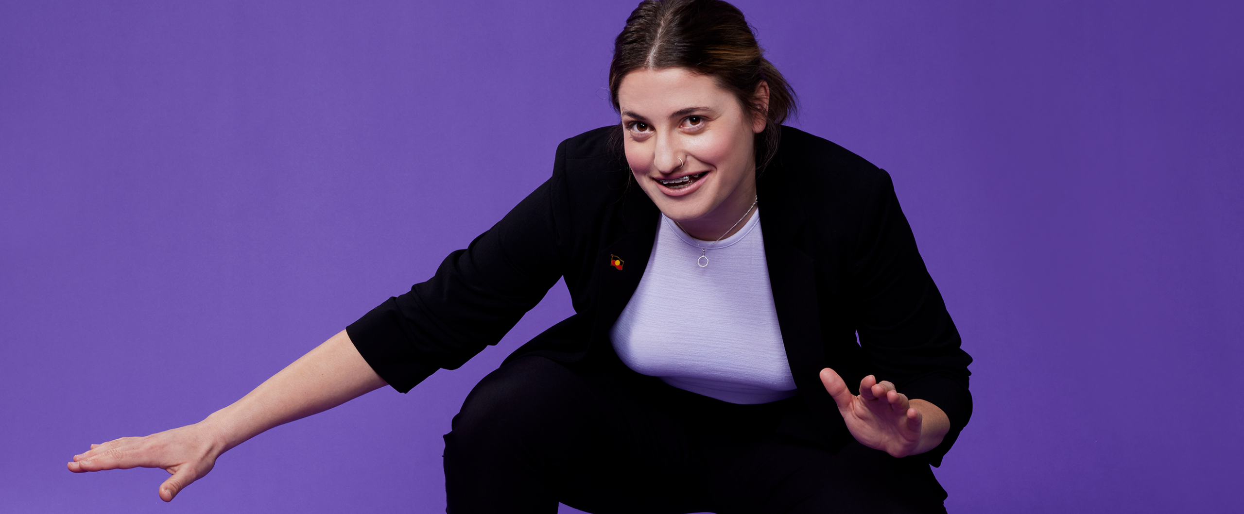 A woman wearing a suit squatting down with both arms out in quite a fun pose in front of a purple background.