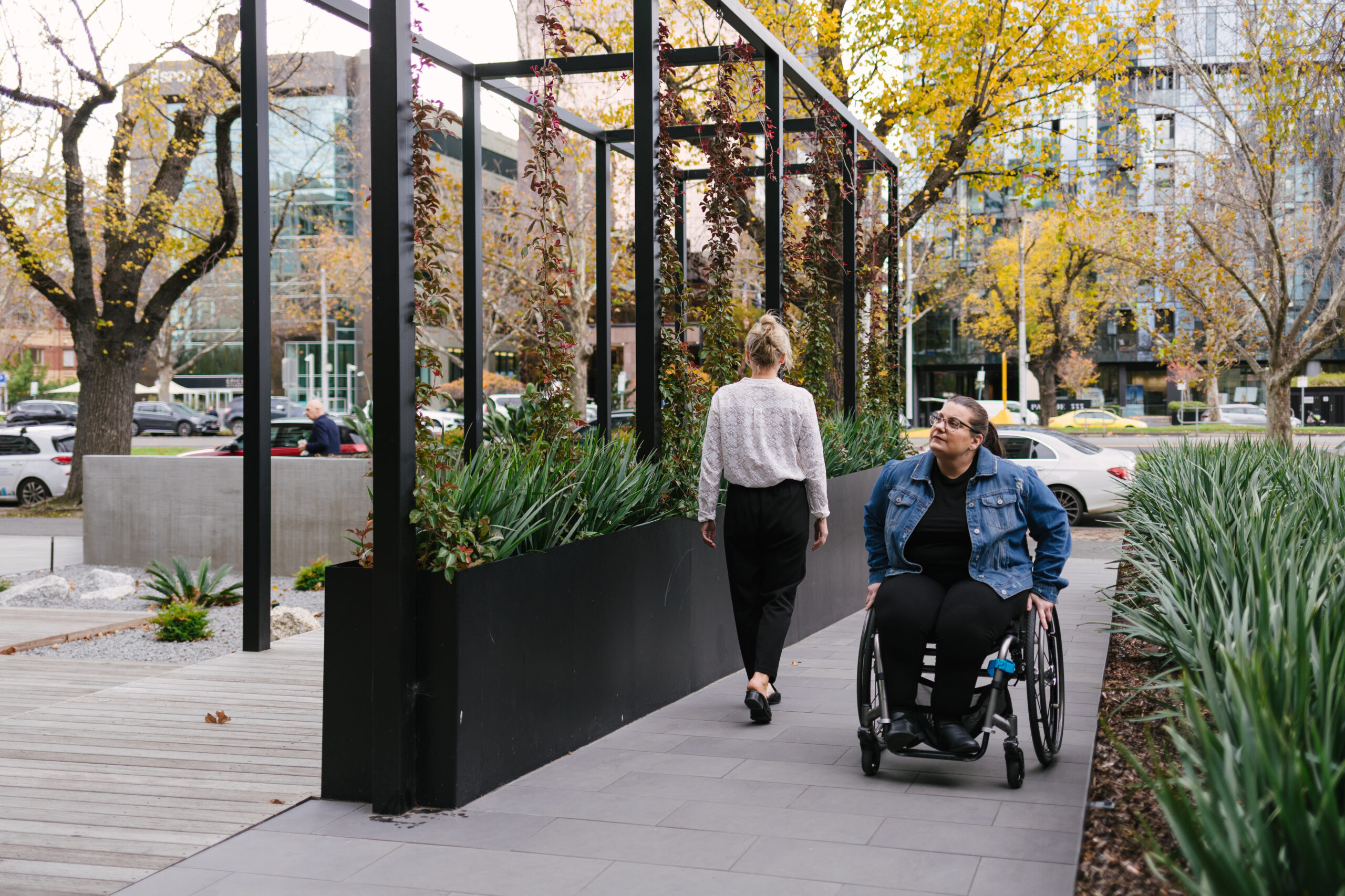 A wheelchair user enjoys the outdoors, facing away from the camera on a sidewalk with lush plants and trees in the surroundings.