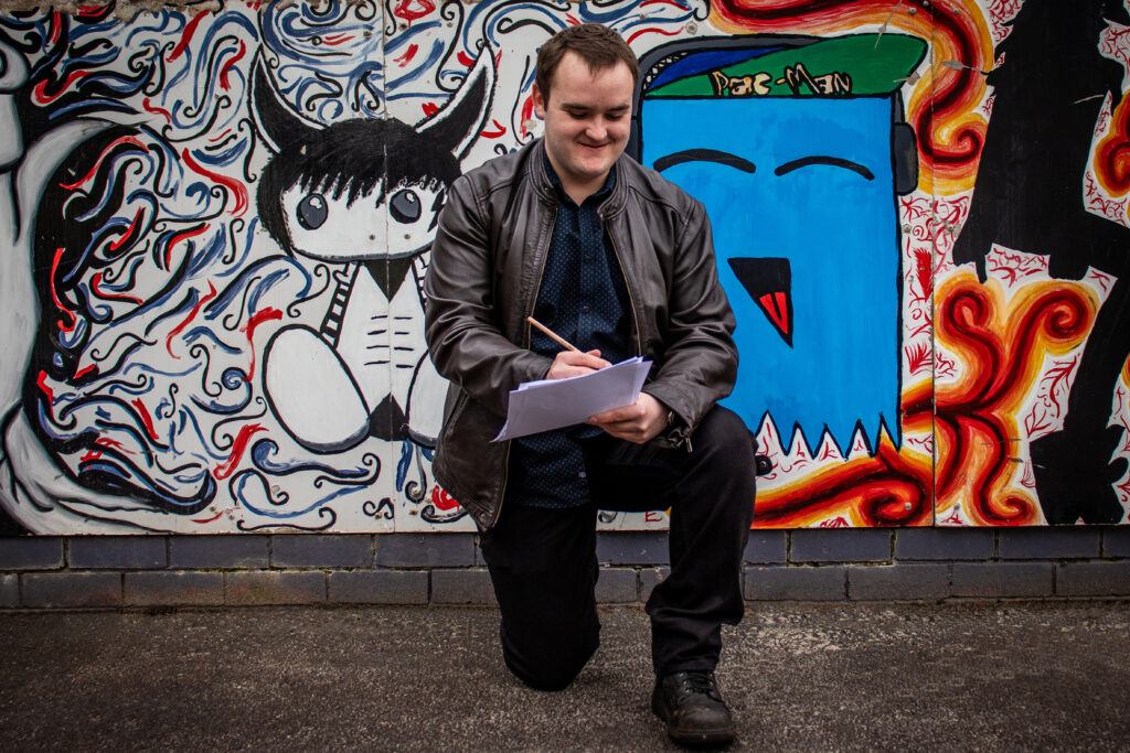 A person kneeling in front of a graffitied wall, smiling and writing on a paper resting on their knee, with a pen in hand.