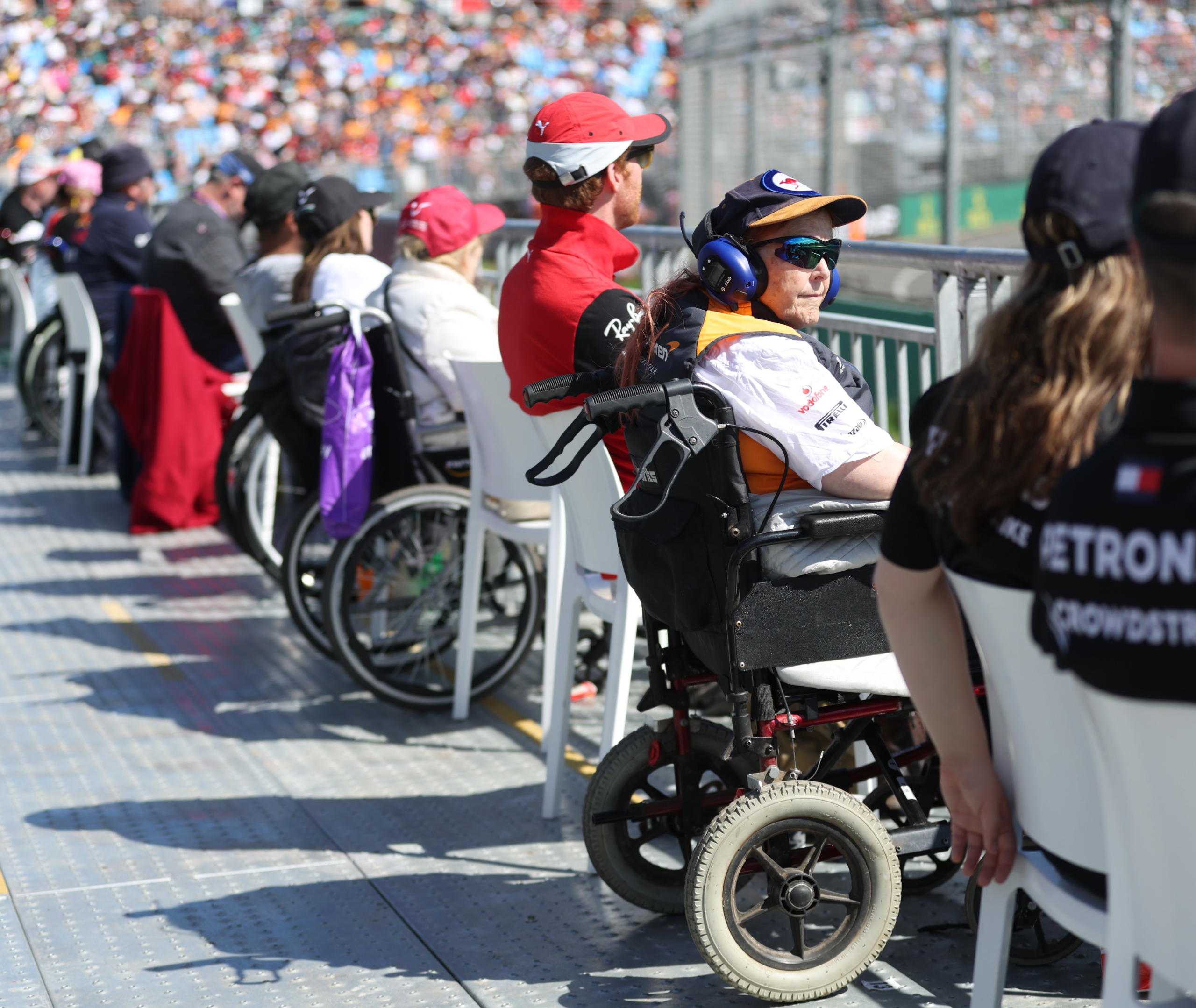 A close-up image of a woman wearing sunglasses and a hat, sitting in a wheelchair with noise-cancellation headphones on, watching the F1 race from an accessible platform.