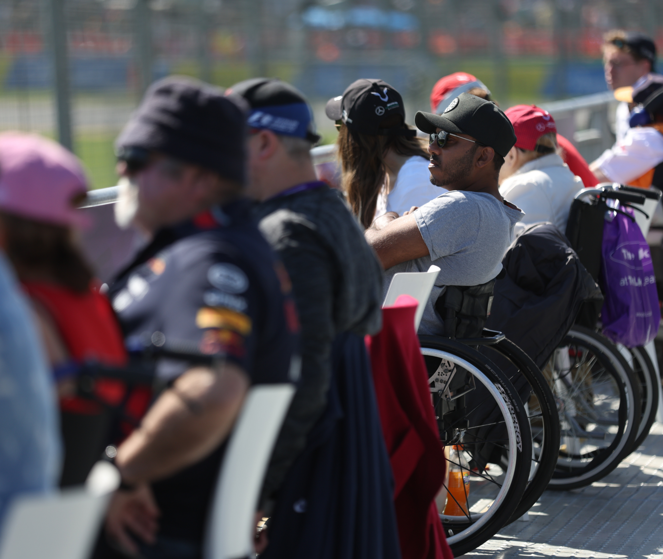 A zoomed-out image of several people watching the Aus GP race from an accessible viewing area. Some of the viewers are in wheelchairs. Most of the audience is dressed in the team colors of F1 racing teams.