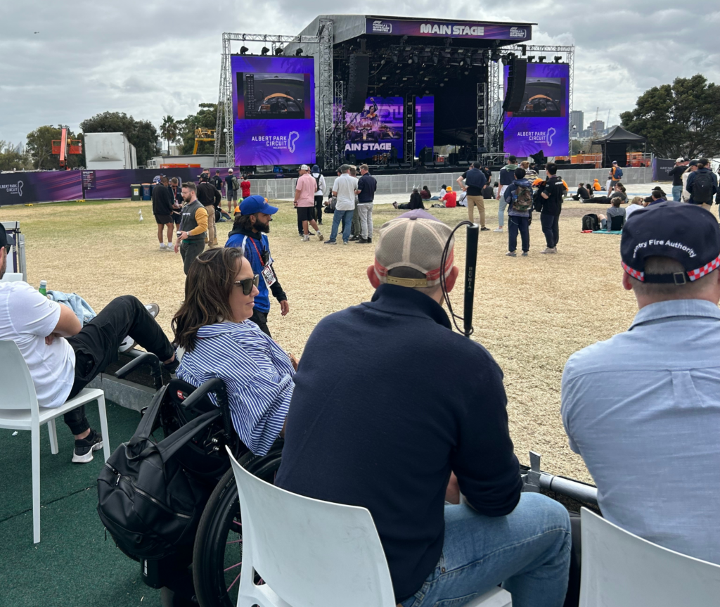 An image of a woman wearing sunglasses in a wheelchair next to a person wearing a hat and holding a walking stick. They are on the accessible viewing platform, with the main stage visible in the background.