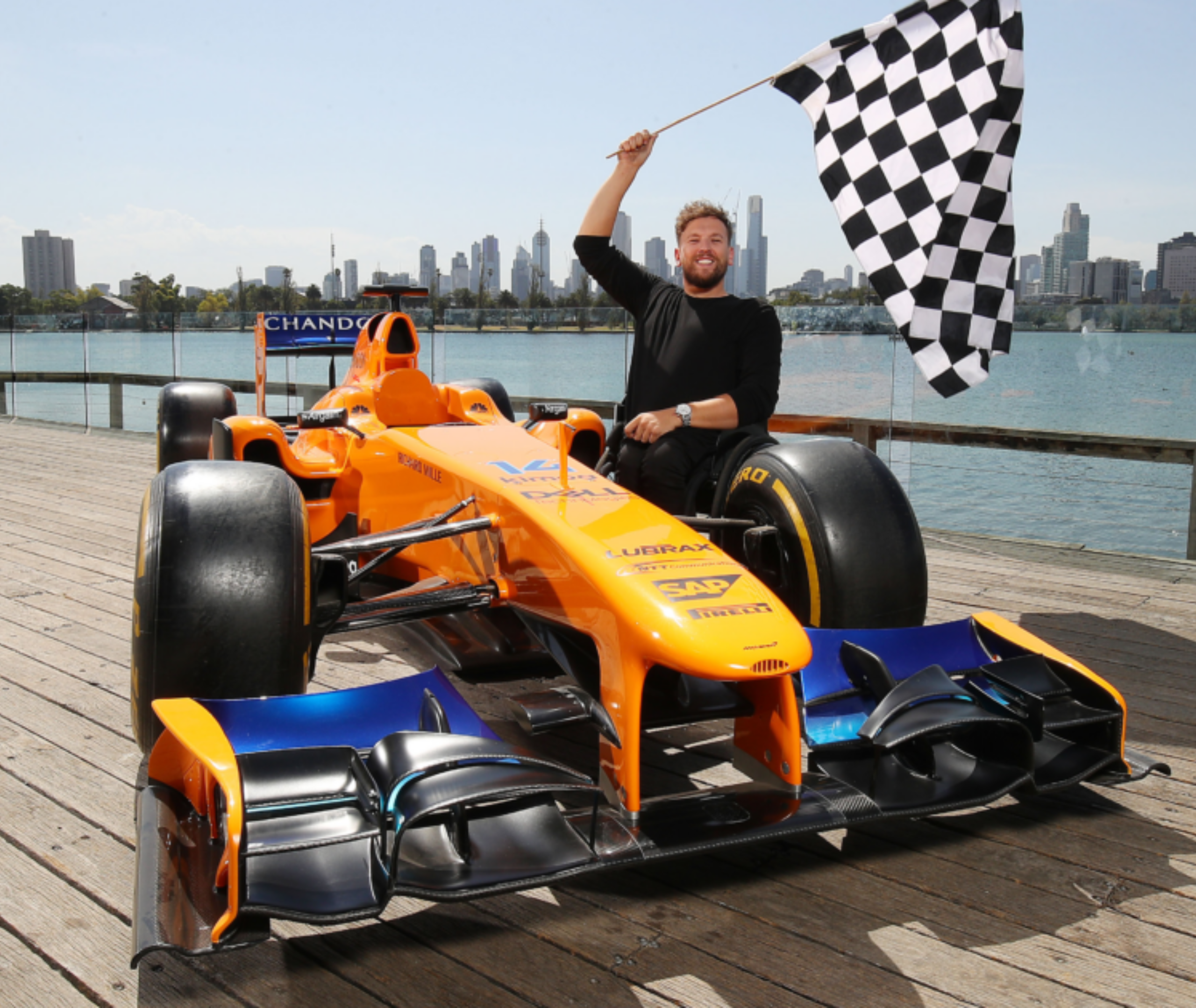 An image of Australian Wheelchair Tennis Champion and former Australian of the Year Dylan Alcott waving the checkered flag next to an F1 car.