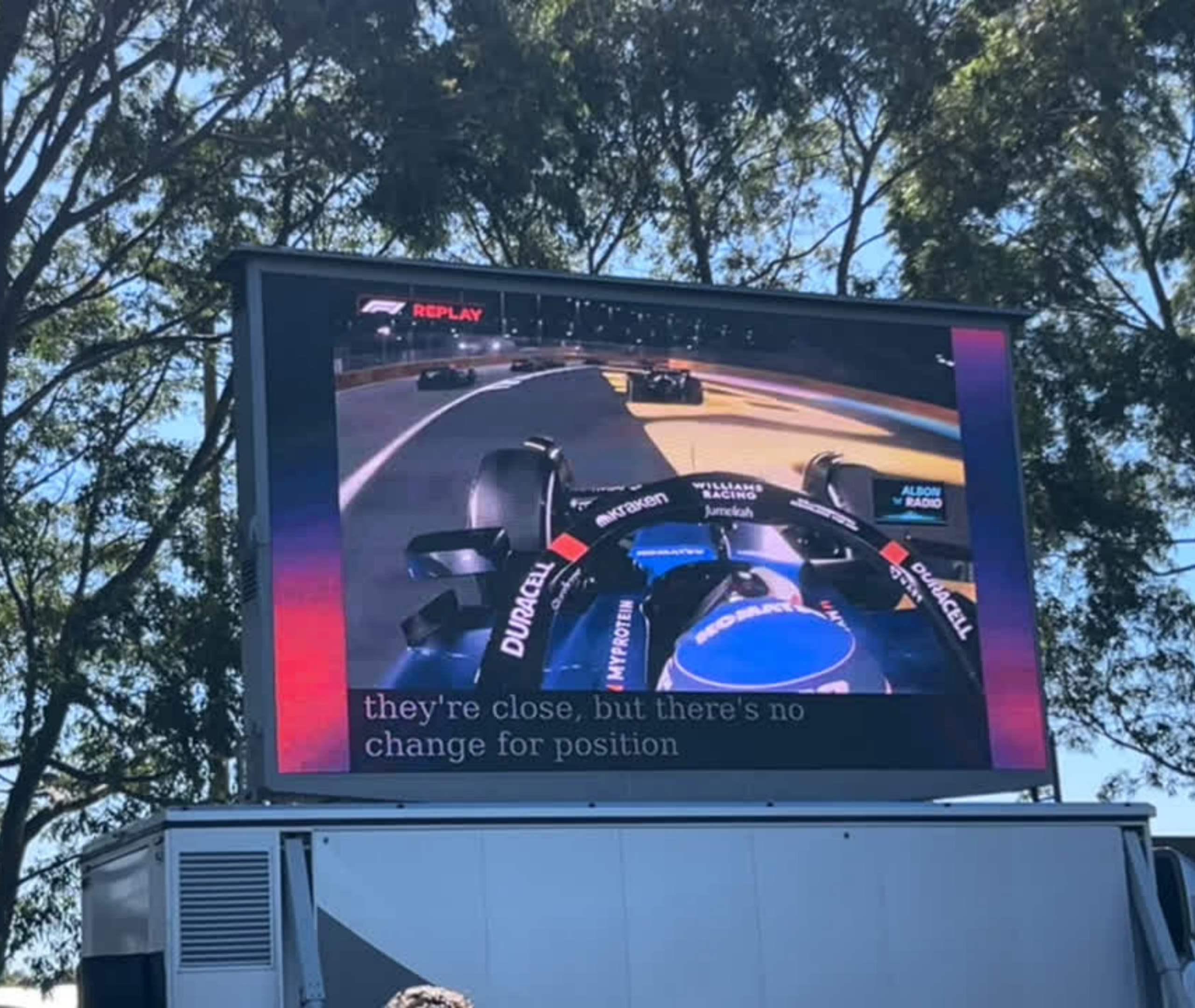 An image of a large screen at the Aus GP showing the race along with closed captions on the screen.