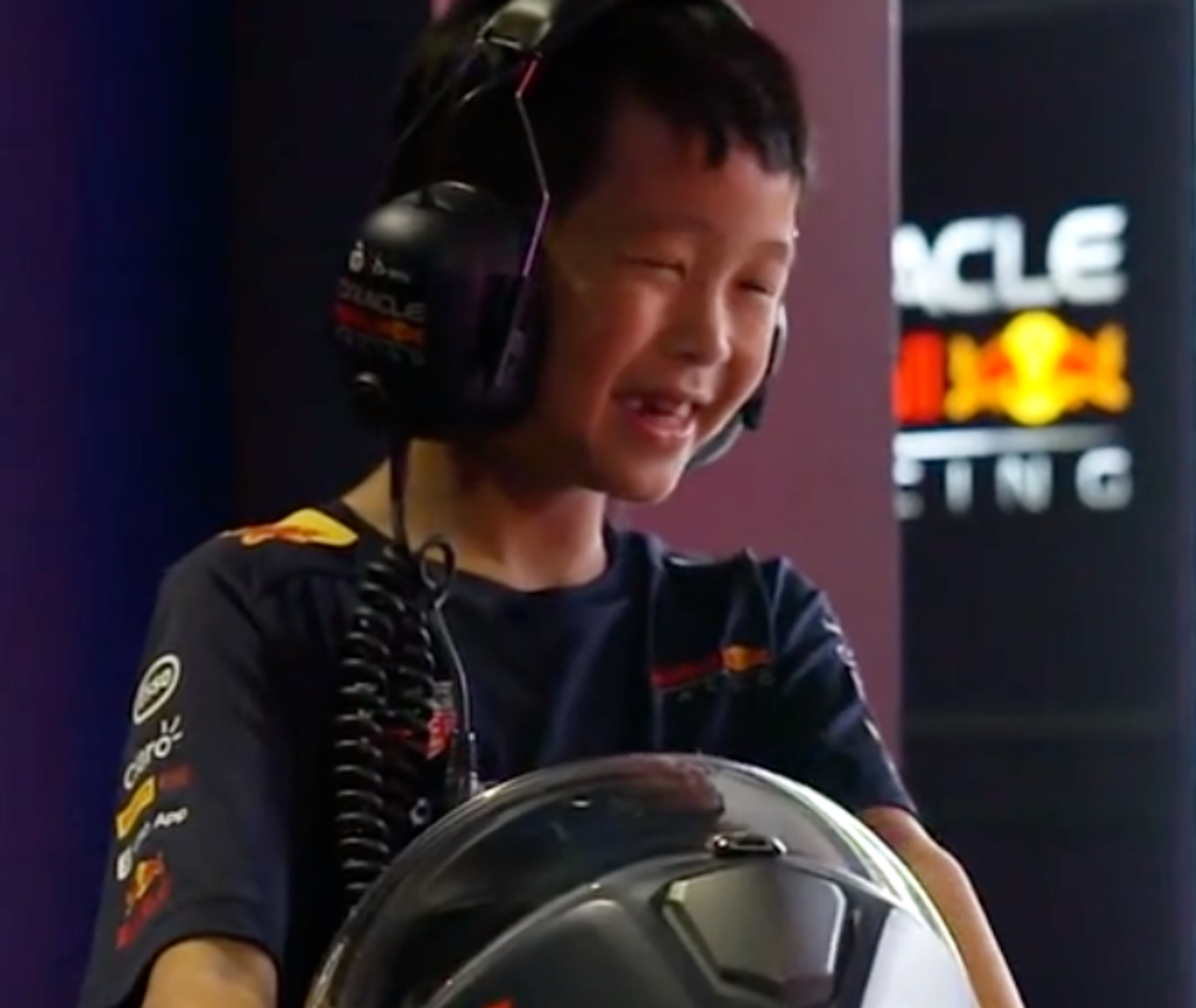 An image of a child holding an F1 racing helmet, wearing noise-cancellation headphones, smiling and looking away from the camera. He is wearing a Red Bull Racing shirt.