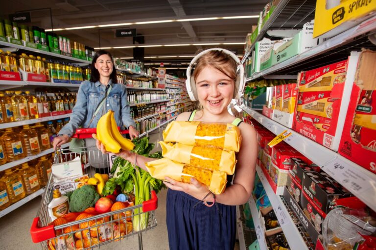 In a Coles shopping center aisle, a mother and daughter are pictured. The mother, in the background, is pushing a shopping cart and smiling at her child. The child, wearing headphones representing the 'Quiet Hour' initiative from Coles, is holding pasta in her left hand and a banana in the other. She is smiling and looking at the camera.
