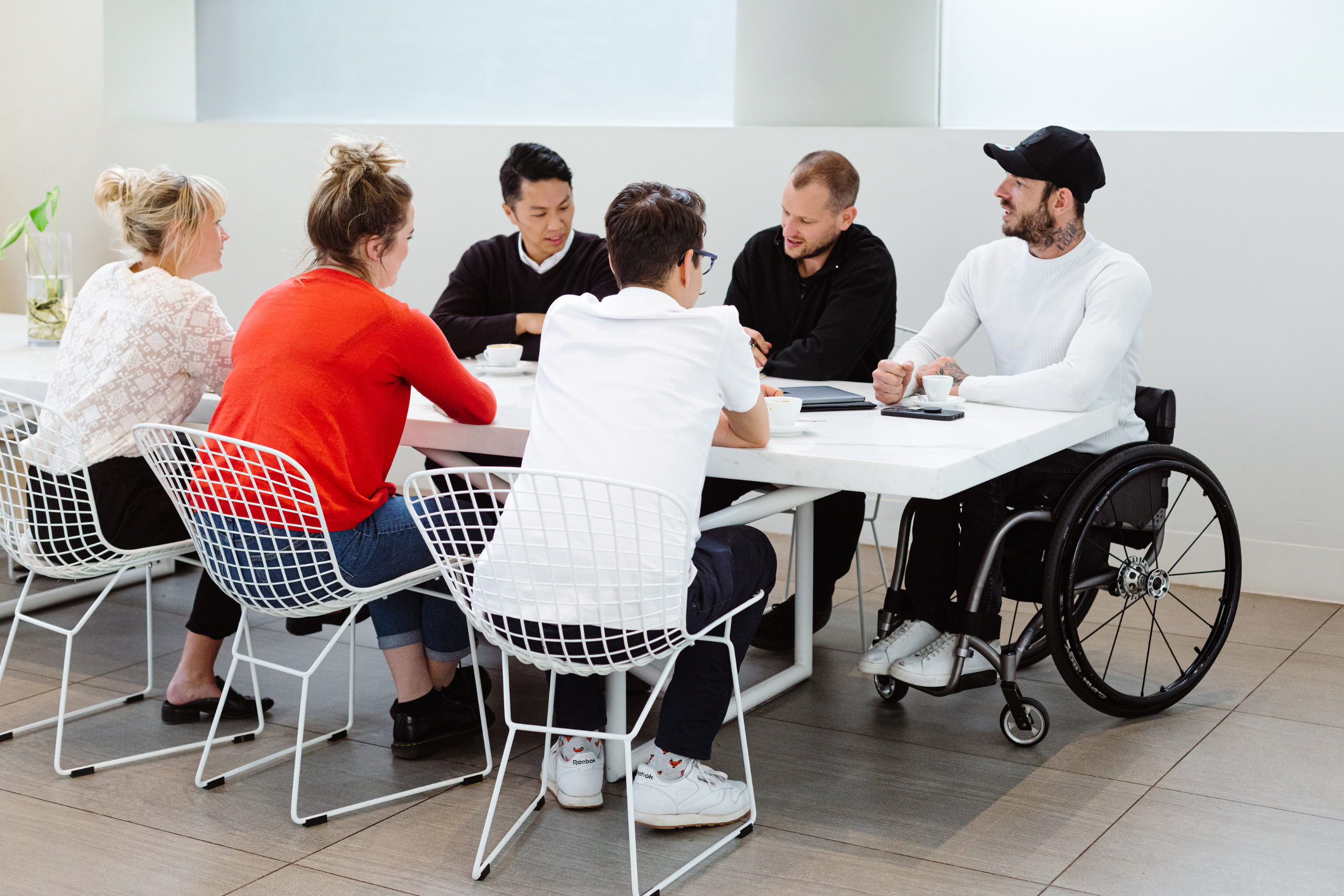 In a meeting room, people are seated around a table, engaged in conversation. At the end of the table, one person is using a wheelchair.