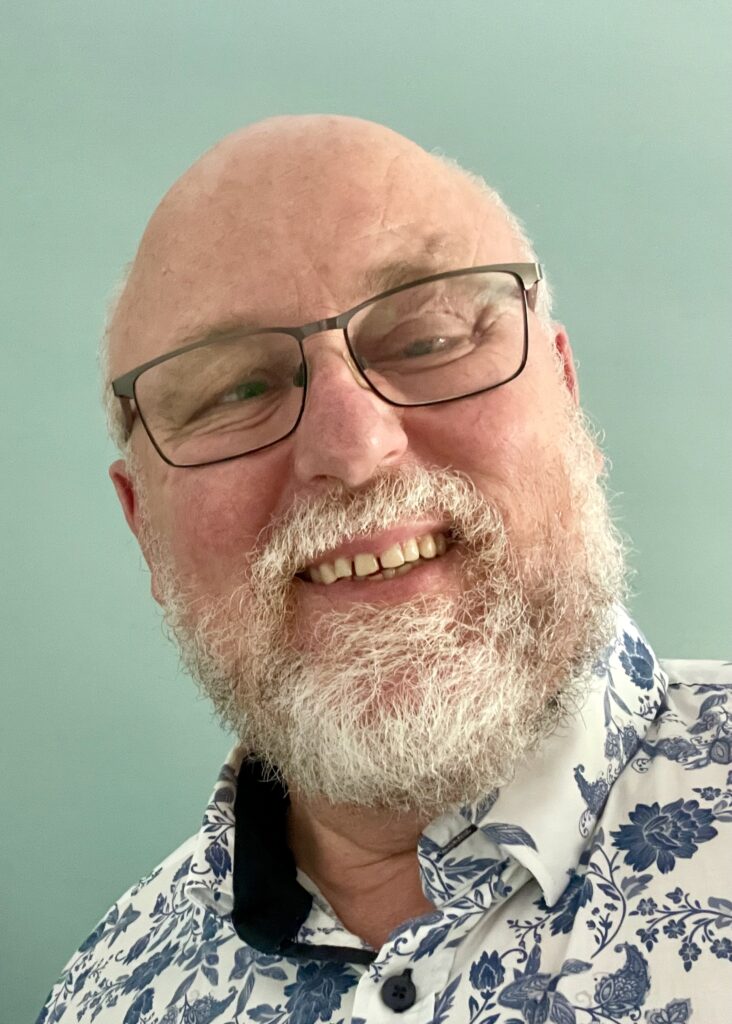 In the selfie, Greg Alchin, a Caucasian male with a white beard, is wearing glasses and smiling as he looks directly at the camera.