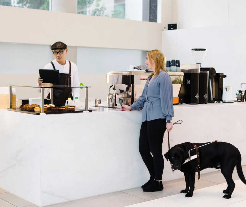 A Caucasian woman with blonde hair, dressed in a blue shirt and black pants, stands at a cafe counter with her guide dog. She is speaking with the barista, a man wearing a white shirt and a black hat. The barista is behind the counter, looking at his screen and typing while conversing with the woman.