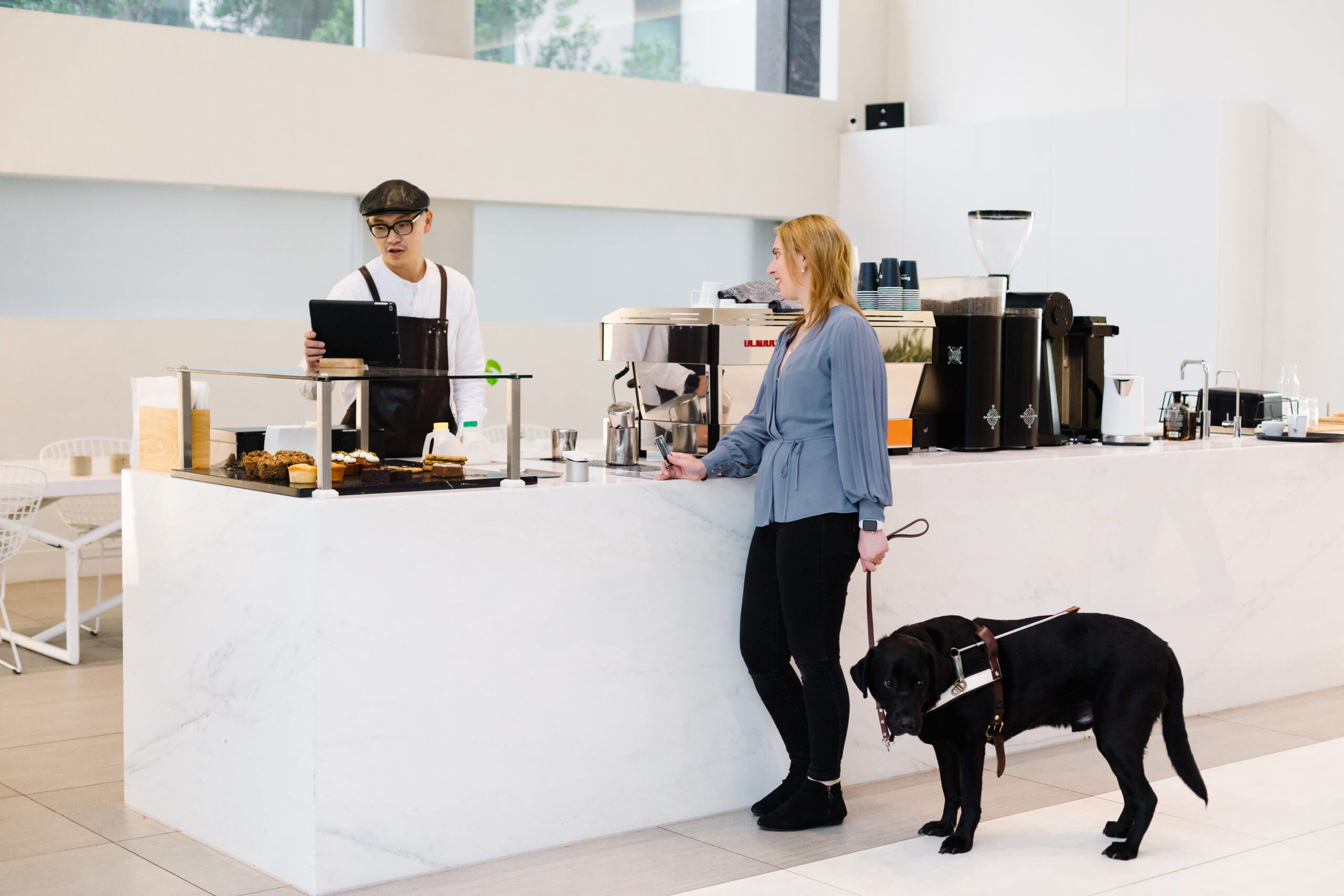 A Caucasian woman with blonde hair, dressed in a blue shirt and black pants, stands at a cafe counter with her guide dog. She is speaking with the barista, a man wearing a white shirt and a black hat. The barista is behind the counter, looking at his screen and typing while conversing with the woman.