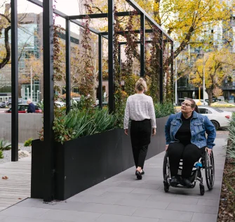 A wheelchair user enjoys the outdoors, facing away from the camera on a sidewalk with lush plants and trees in the surroundings.