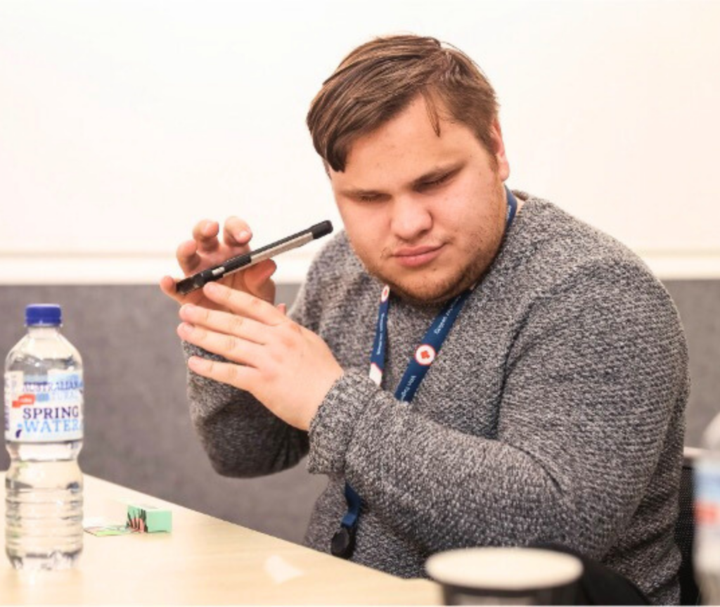 A Caucasian male with brown hair is wearing a grey jacket. He is seated on a chair with a table in front of him. In the image, he is using a screen reader on his mobile phone, holding the device close to his ear as he listens.