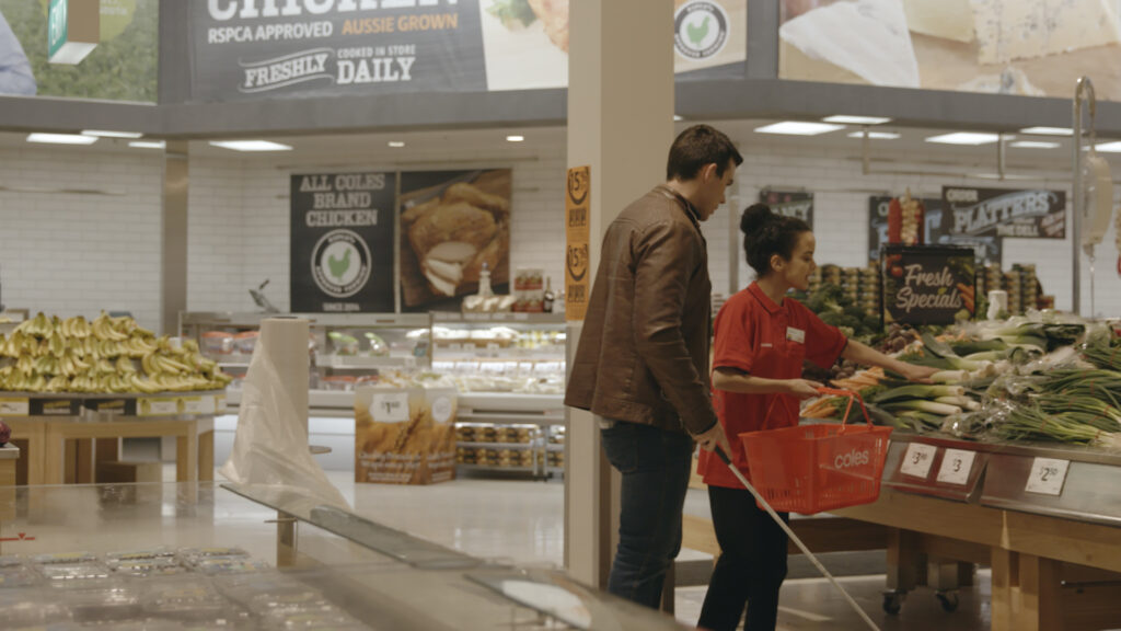 A person wearing a brown jacket and using a walking stick is accompanied by a Coles staff member holding a shopping basket. They are shopping together in the vegetables section.