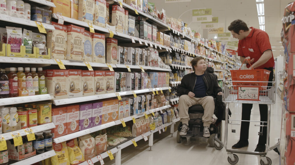 A person on a wheelchair is interacting with a Coles staff member in a Coles store aisle. The aisle is filled with shelves stocked with various products.