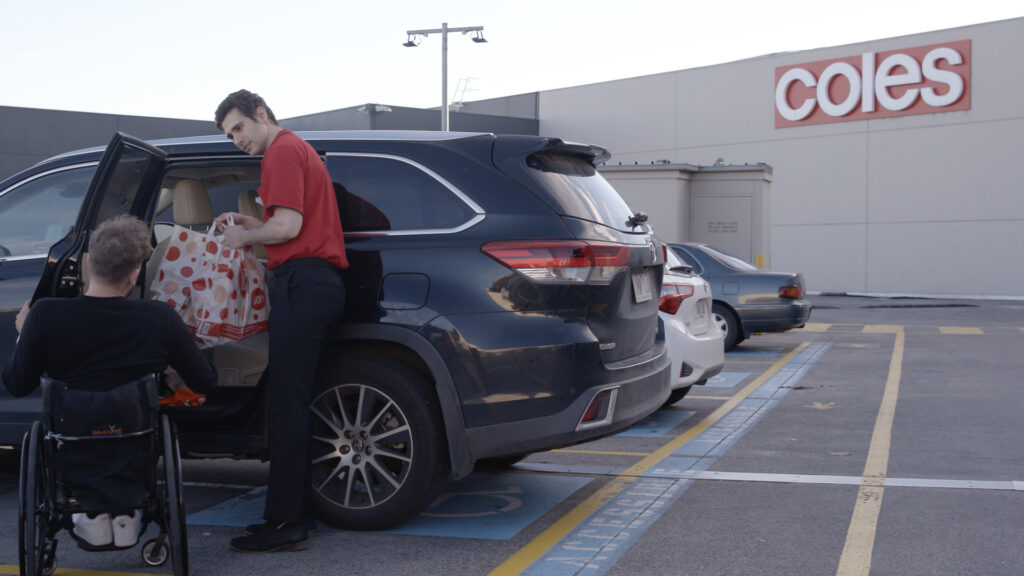 A Coles staff member is helping a customer who uses a wheelchair. The staff member is assisting by putting shopping bags into the customer's black car.