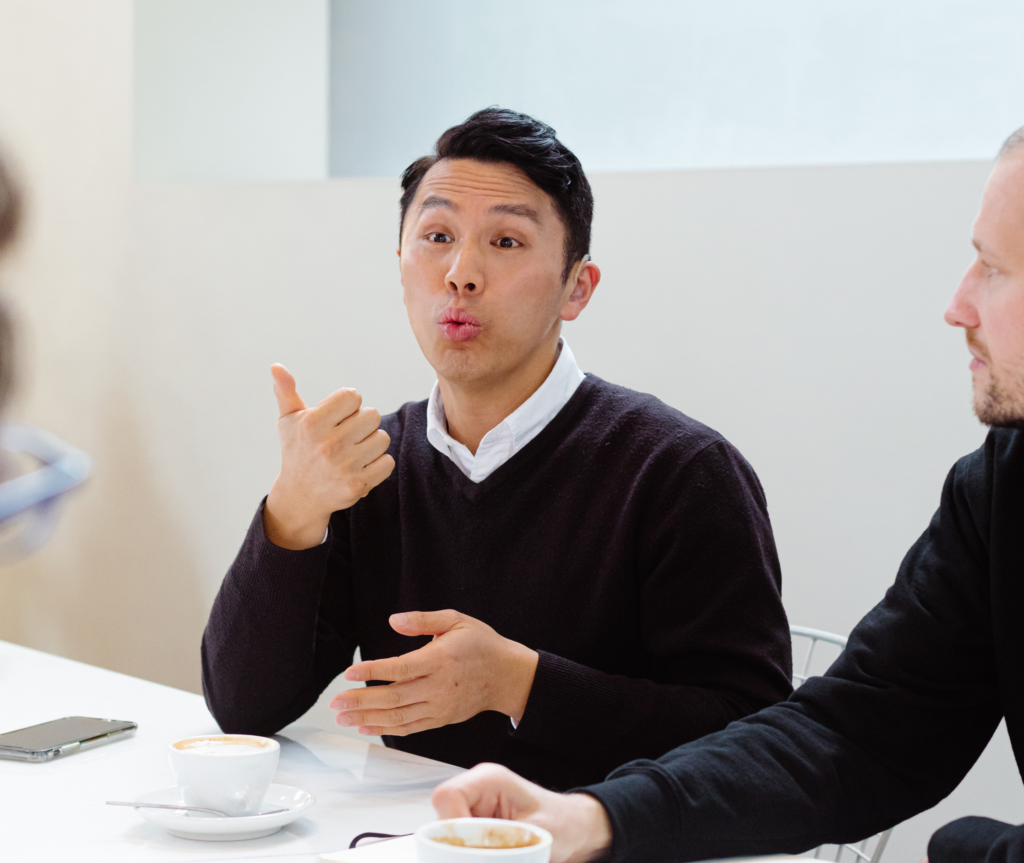 Elvin, a Caucasian man with dark hair, is wearing a black jacket and sitting at a table with a cup of coffee in front of him. He is wearing a hearing aid and is using Auslan sign language to communicate with others seated around the table.