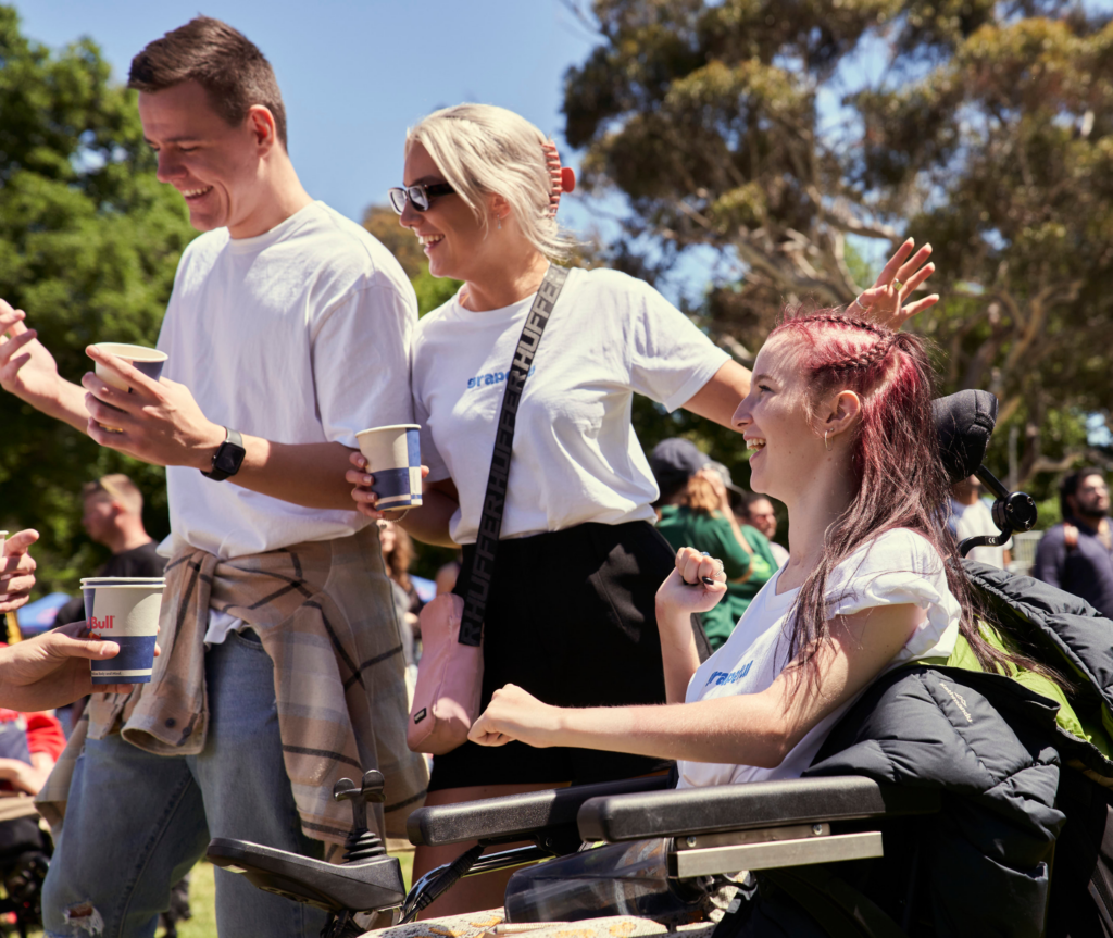 A person in a wheelchair is smiling as they make their way into the festival, accompanied by others who are also smiling. In the background, additional festival-goers are visible, contributing to the vibrant atmosphere.