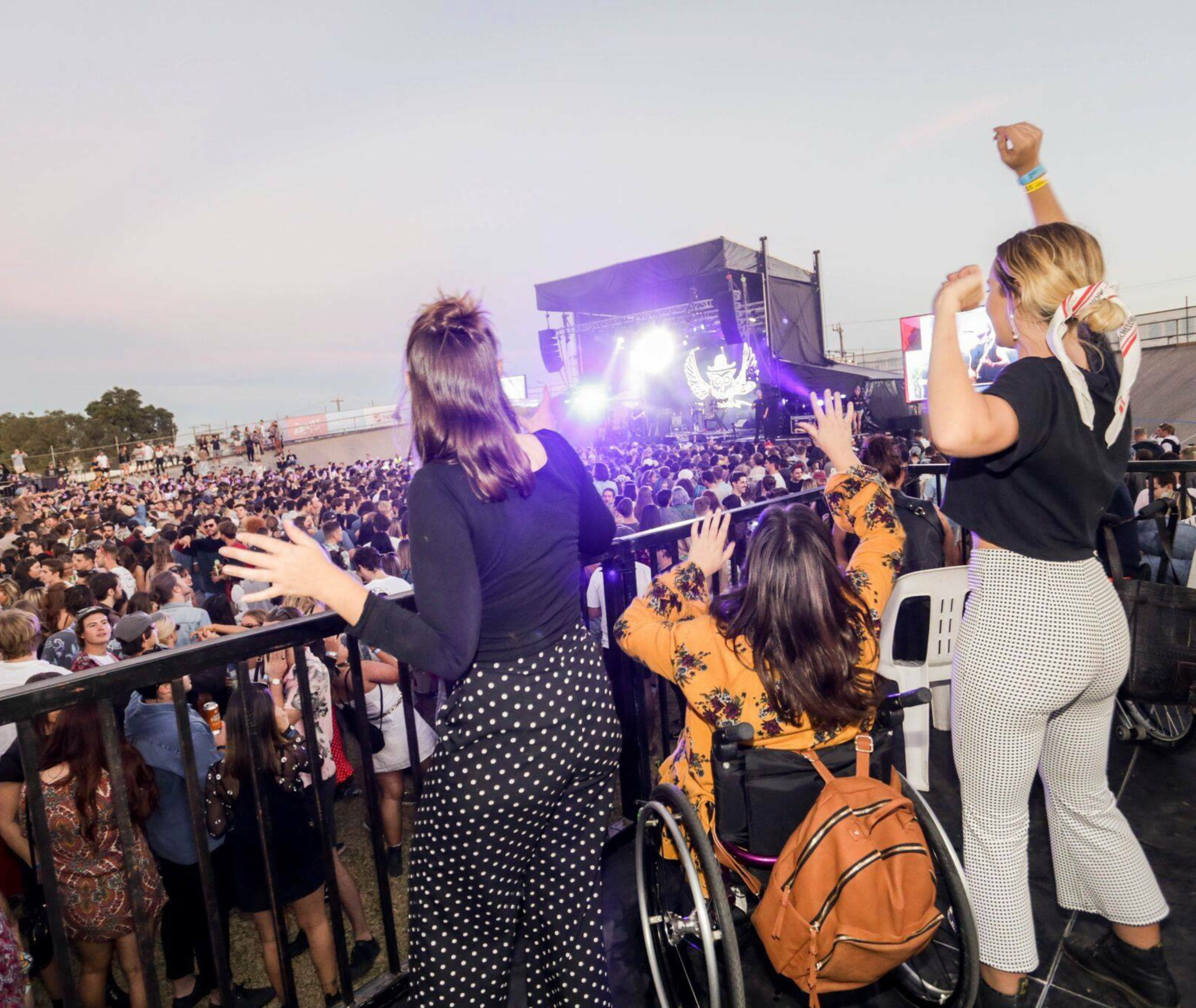 A person in a wheelchair is positioned on an accessible ramp at a music festival, facing toward the stage. The stage is visible in the background, illuminated by various lights. Around the ramp, festival-goers are dancing and enjoying the performance, adding to the lively atmosphere.