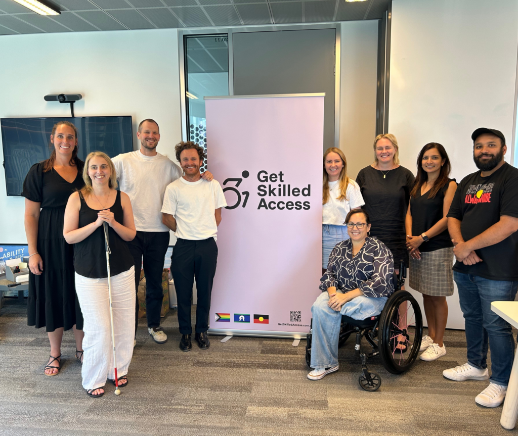 Group of Get Skilled Access staff smiling for the camera in a meeting room. The GSA pull-up banner is positioned in the center of the group.