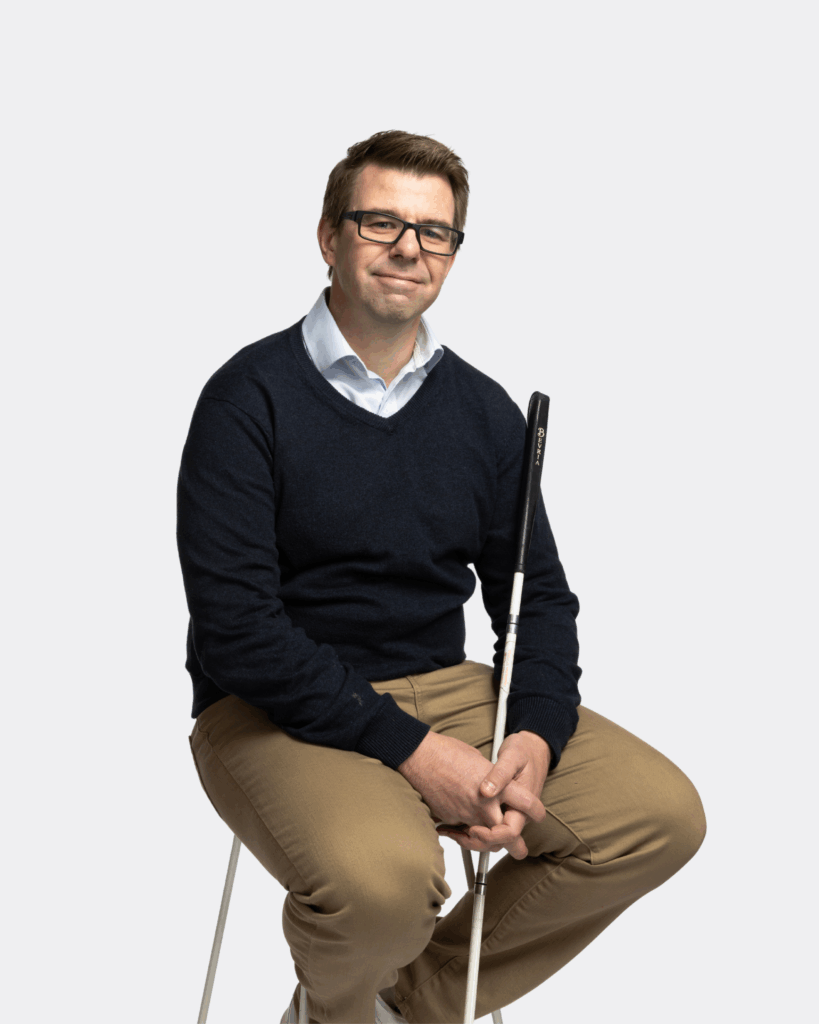 Travis Zimmer, a person using white cane is wearing black jumper and brown trousers. He is sitting on a chair in an angle and looking at the camera and smiling.
