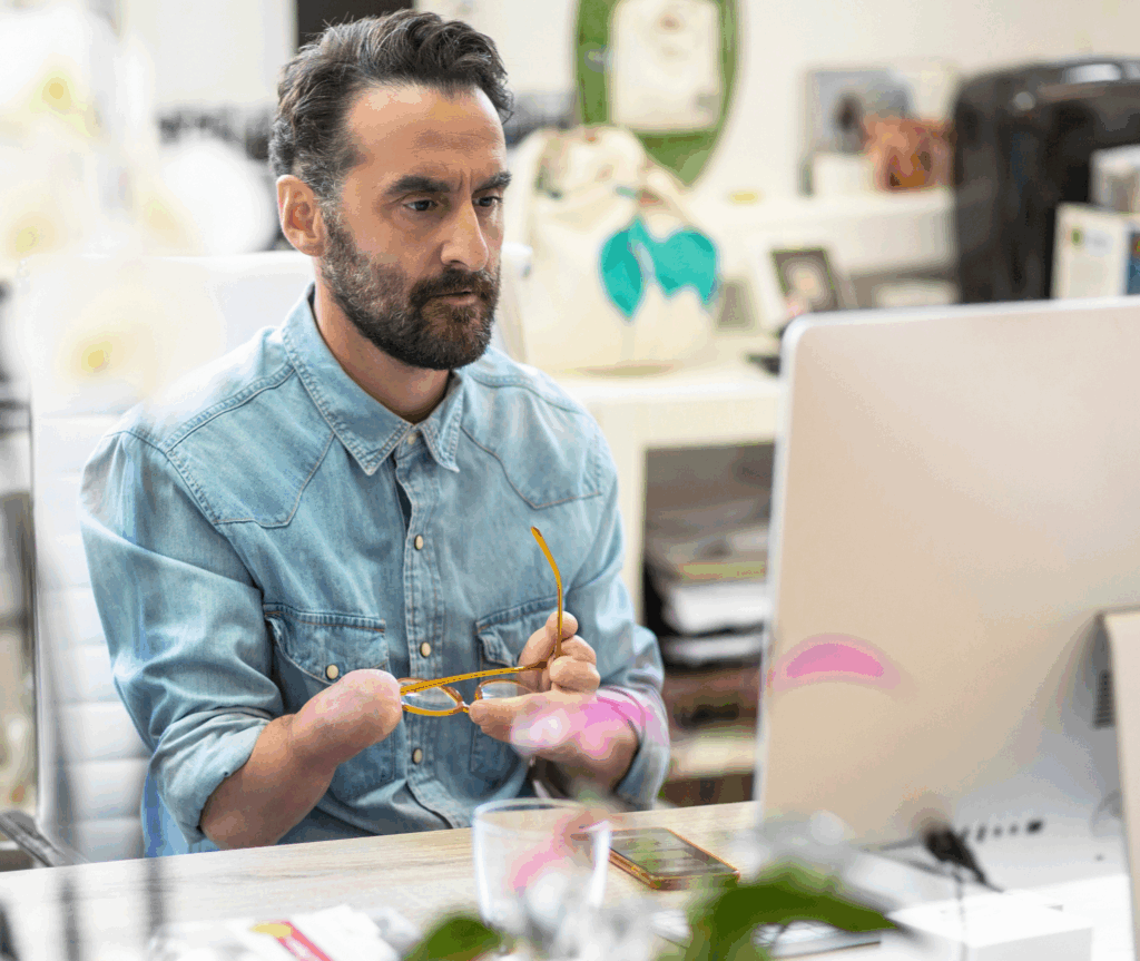 Man with a limb difference below the left wrist sits at a desk, holding glasses and looking at a computer screen.