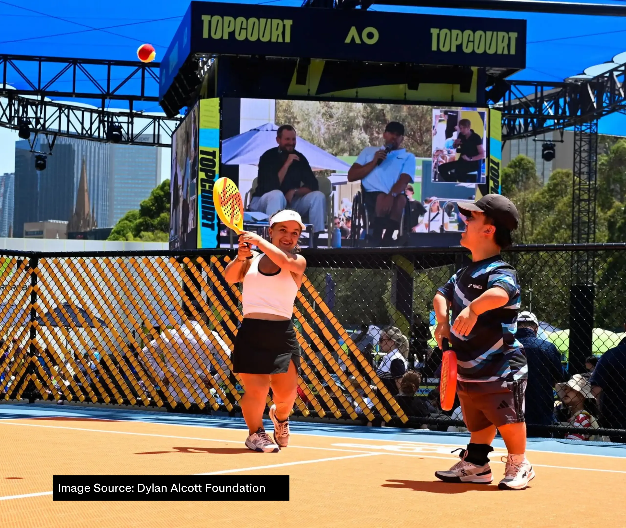 At AO 2025, a large screen shows Dylan Alcott and Zack Alcott speaking with an Auslan interpreter beside them under the heading "Top Court". In the foreground, two short statured tennis players are playing on court.