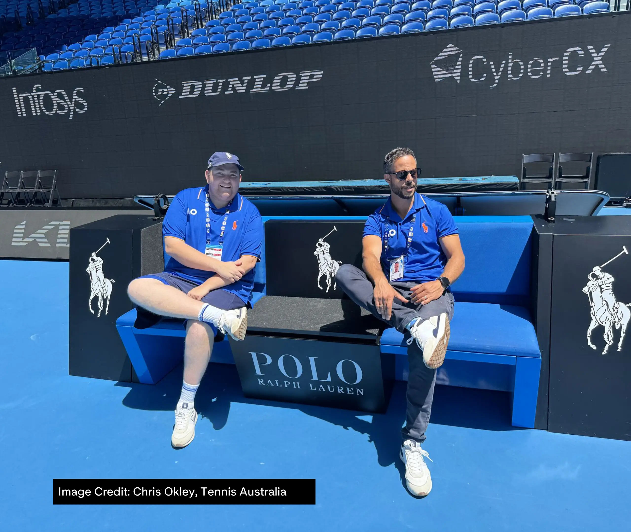 Kerry Tavrou and Chris Okely from Tennis Australia sit on a bench on the tennis court at AO 2025, both wearing blue AO polos.