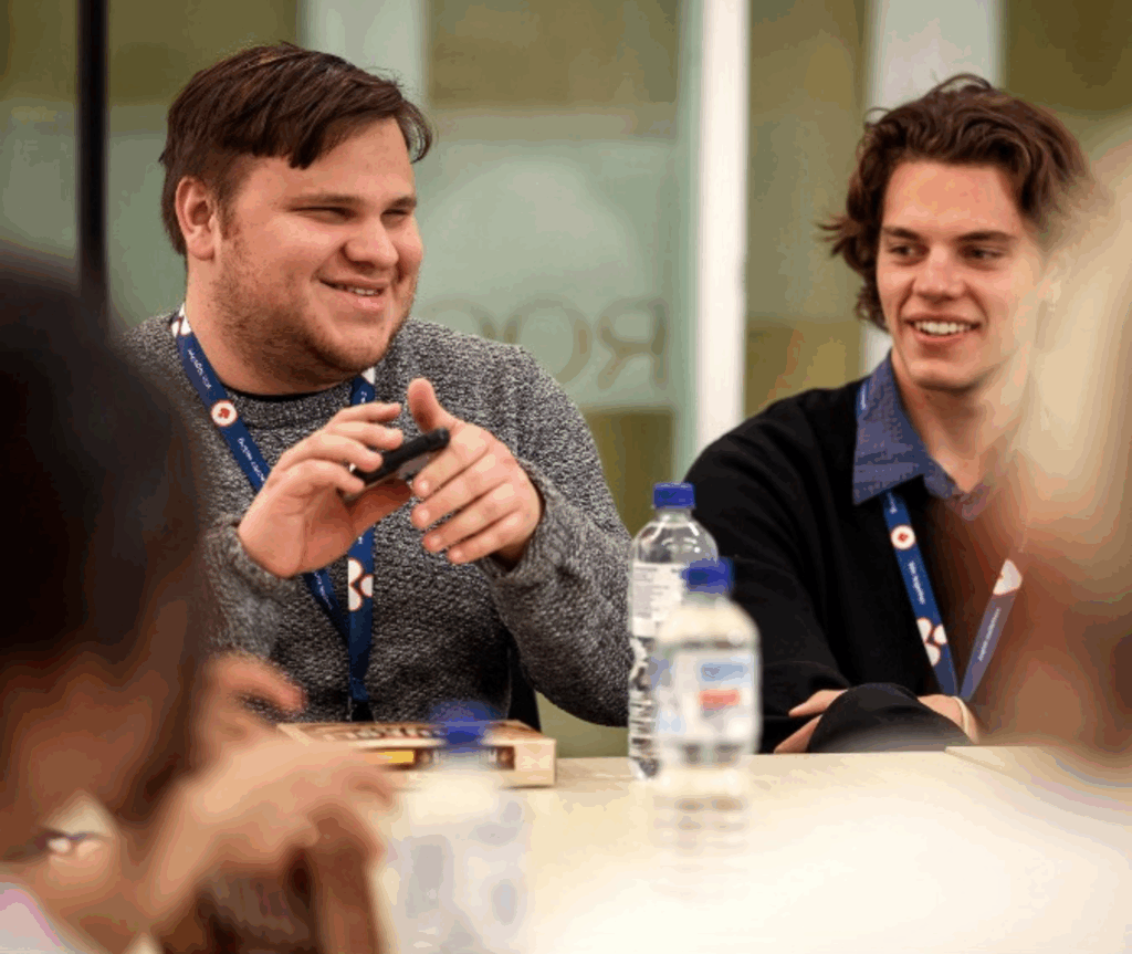 Nathan, a GSA associate who is blind, is testing a product, holding a phone and listening to the audio description. He is wearing a grey jumper and smiling while speaking with someone, while another person observes.