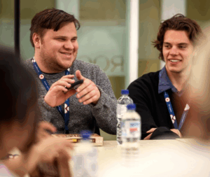 Nathan, a GSA associate who is blind, is testing a product, holding a phone and listening to the audio description. He is wearing a grey jumper and smiling while speaking with someone, while another person observes.