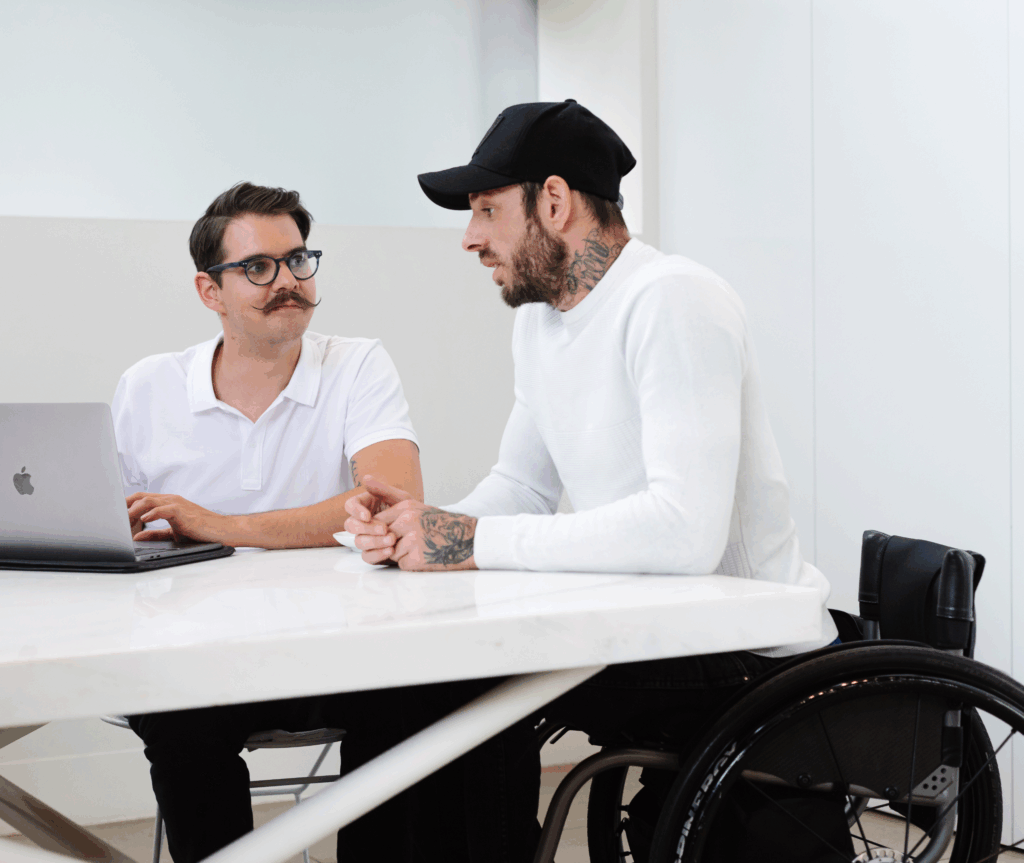 A man on wheelchair next to another person who has a laptop in front of him. Both are engaged in a conversation.
