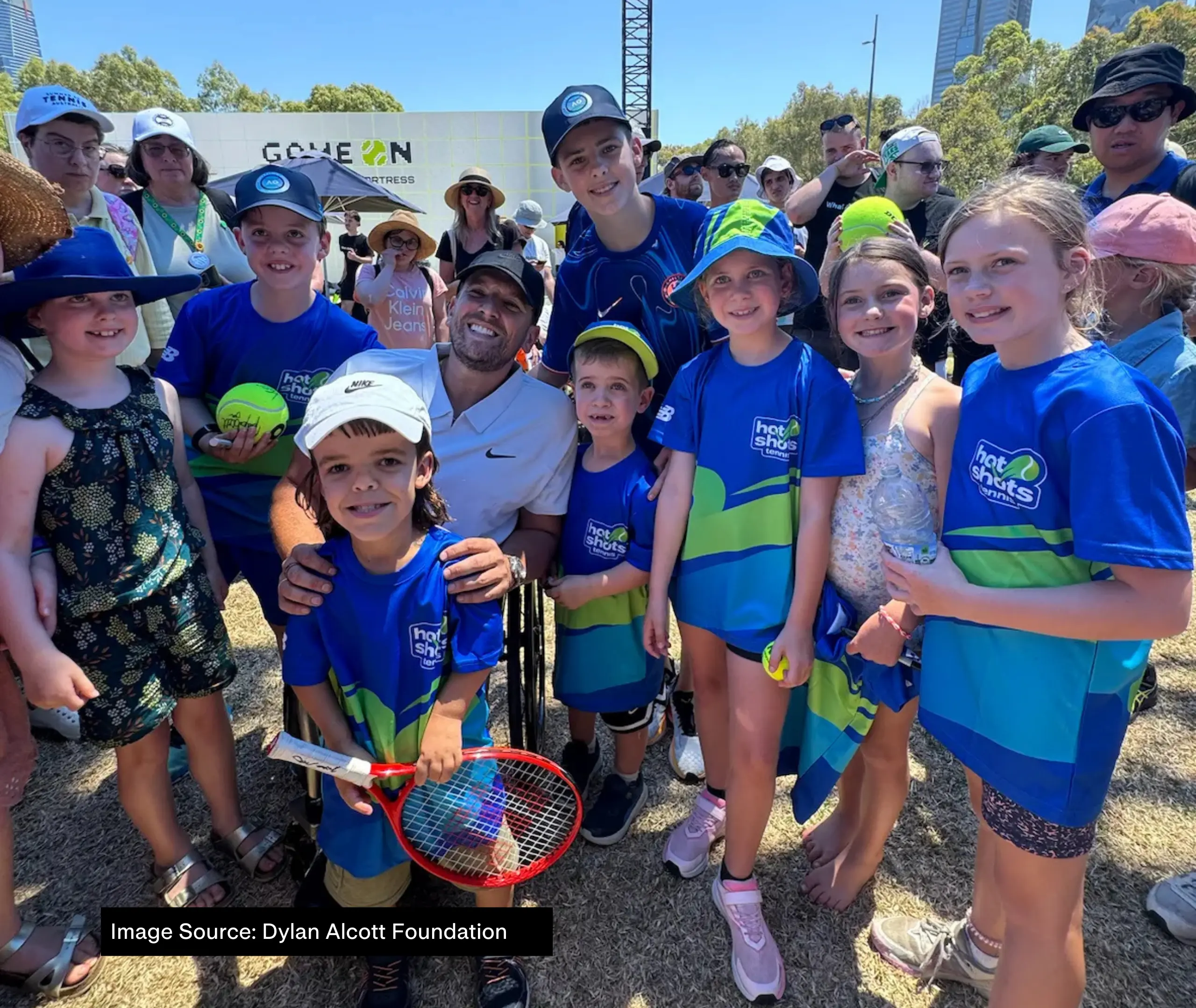 A group of young people wearing Tennis Hot Shots program blue polos are gathered around Dylan Alcott for a group photo. They are all standing close together, looking at the camera.