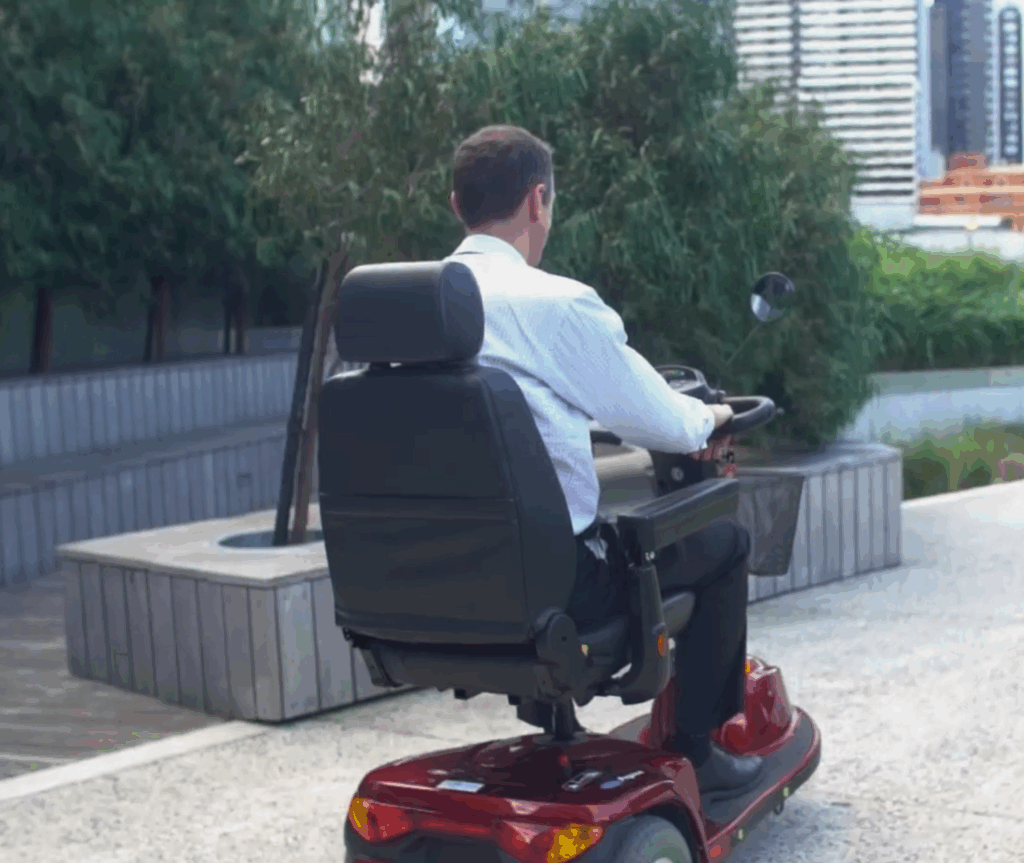 Man using a mobility scooter outdoors, travelling along a paved path with greenery and buildings in the background.