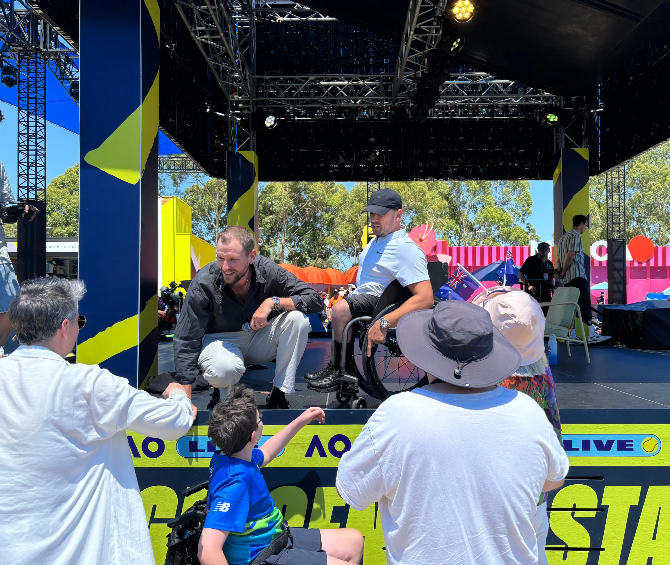 Zack Alcott is on the elevated Top Court stage at AO 2025, bending down to speak with someone at ground level. Next to him, Dylan Alcott, seated in a wheelchair, is approaching a young person who is reaching out to shake hands and engage with him. Several other people are gathered around the front of the stage, creating a lively and interactive atmosphere.