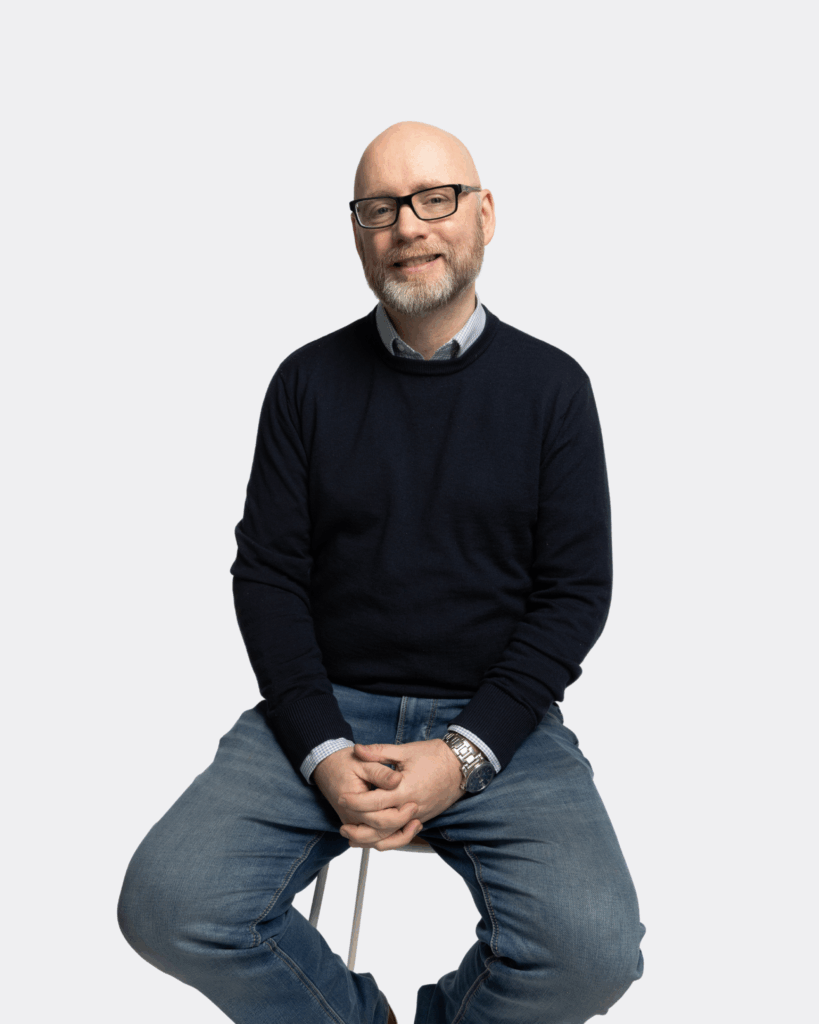 Paul Knox, GSA Bid Manager, is wearing a black jumper and blue jeans, seated on a chair, looking at the camera and smiling.