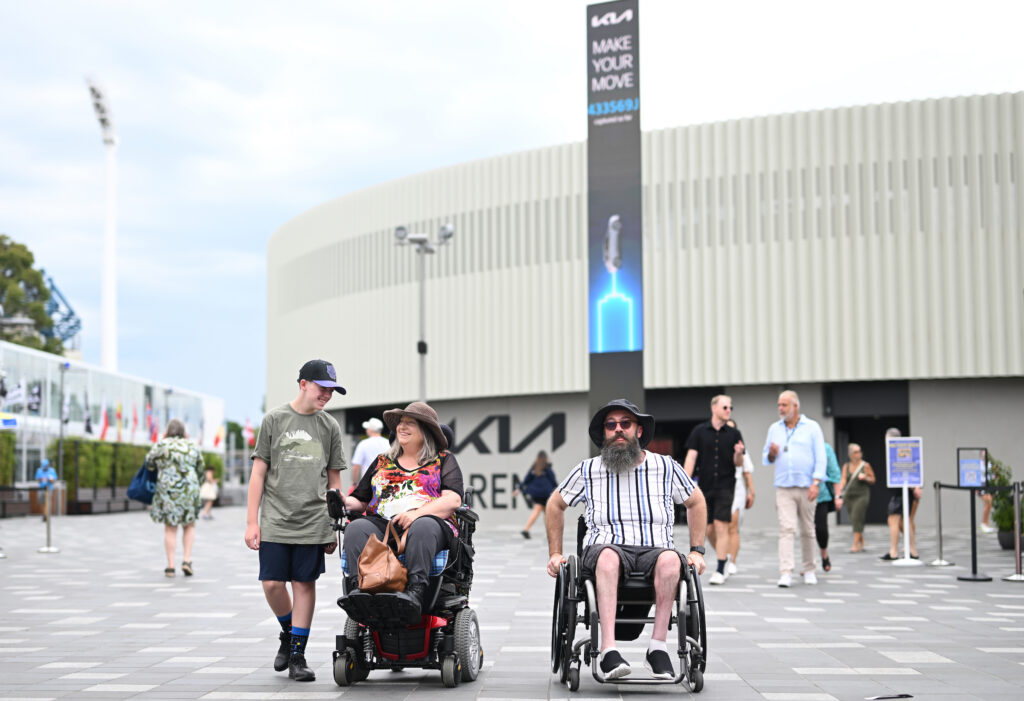Two people on wheelchair accompanied by another person walking beside them.