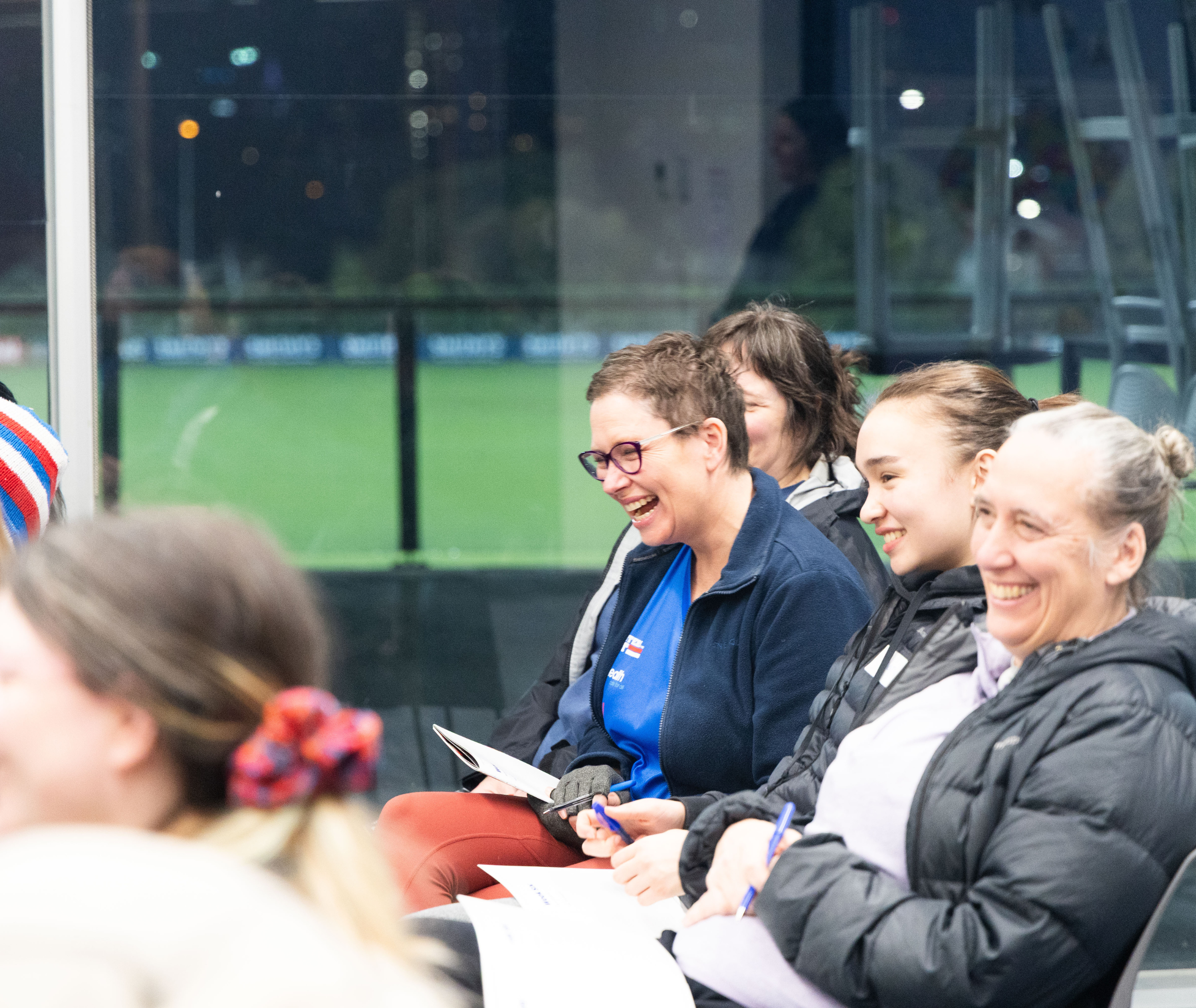 A group of people sitting together and laughing during an indoor workshop, holding papers and pens, with a sports field visible through the glass wall behind them.