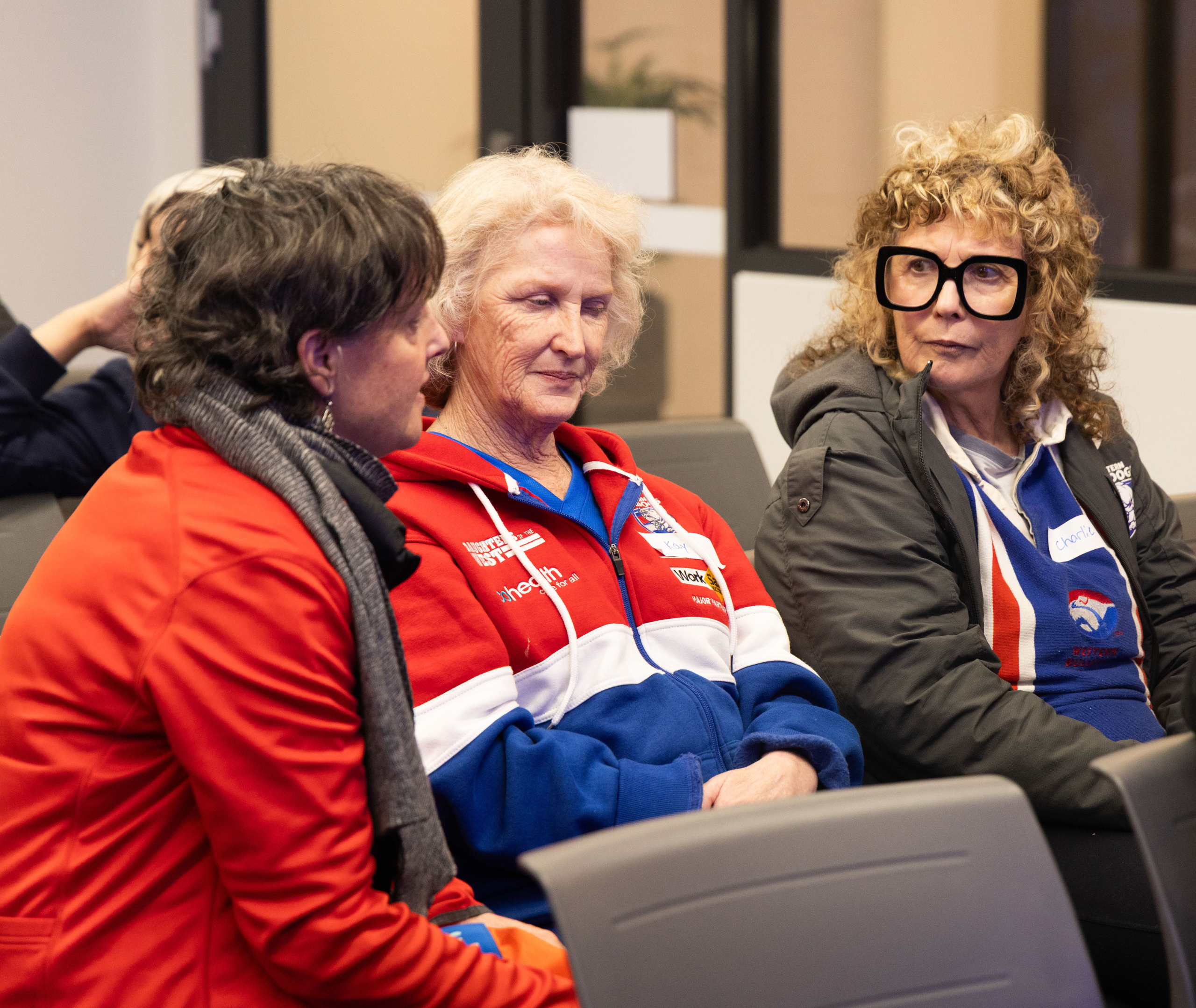 Three women in red, white, and blue jackets talking together in an indoor seating area before a session begins.