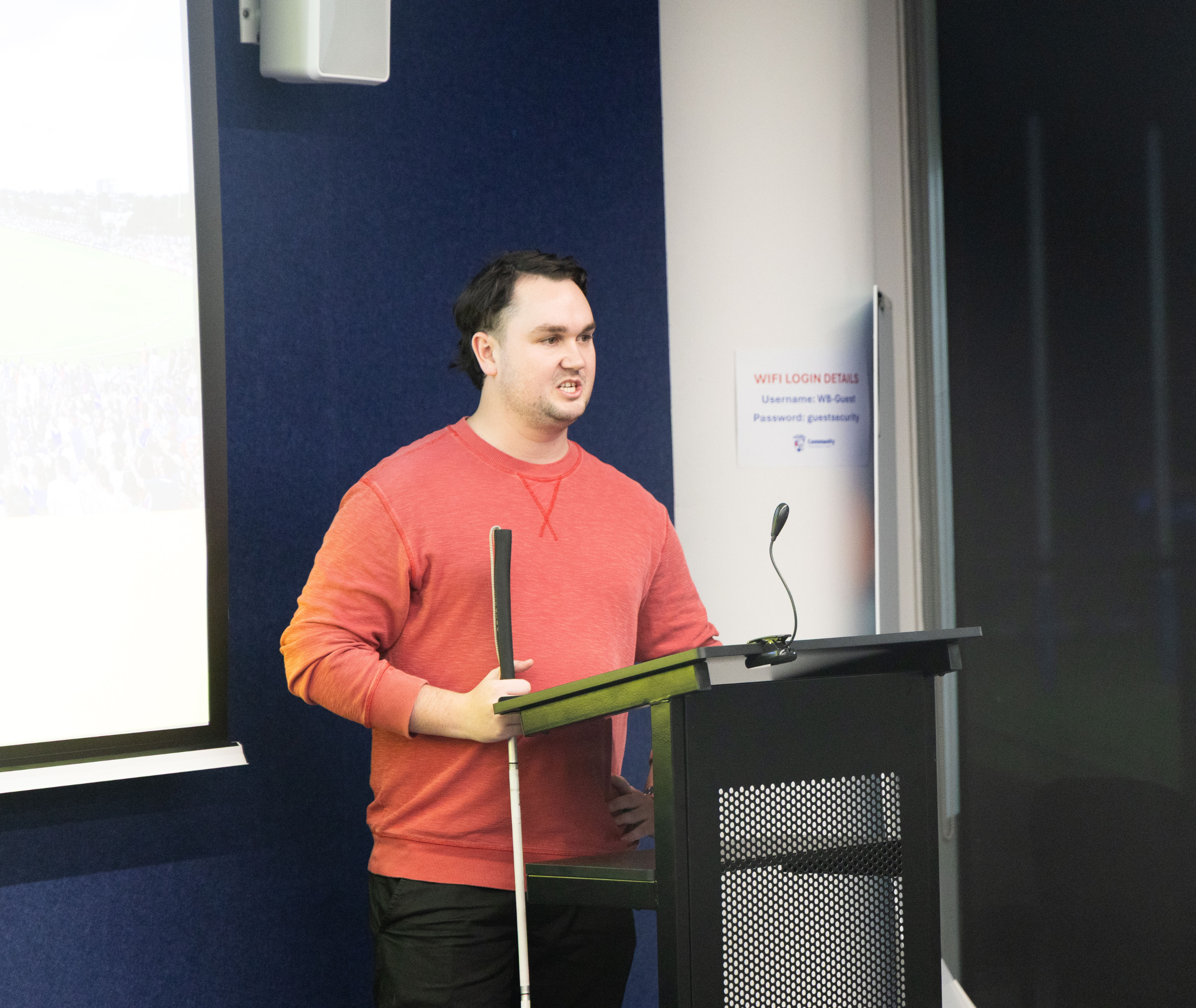 Sport4All State Manager Johnny is wearing a red jumper speaking at a lectern with a white cane in hand, presenting in front of a screen at an indoor session.