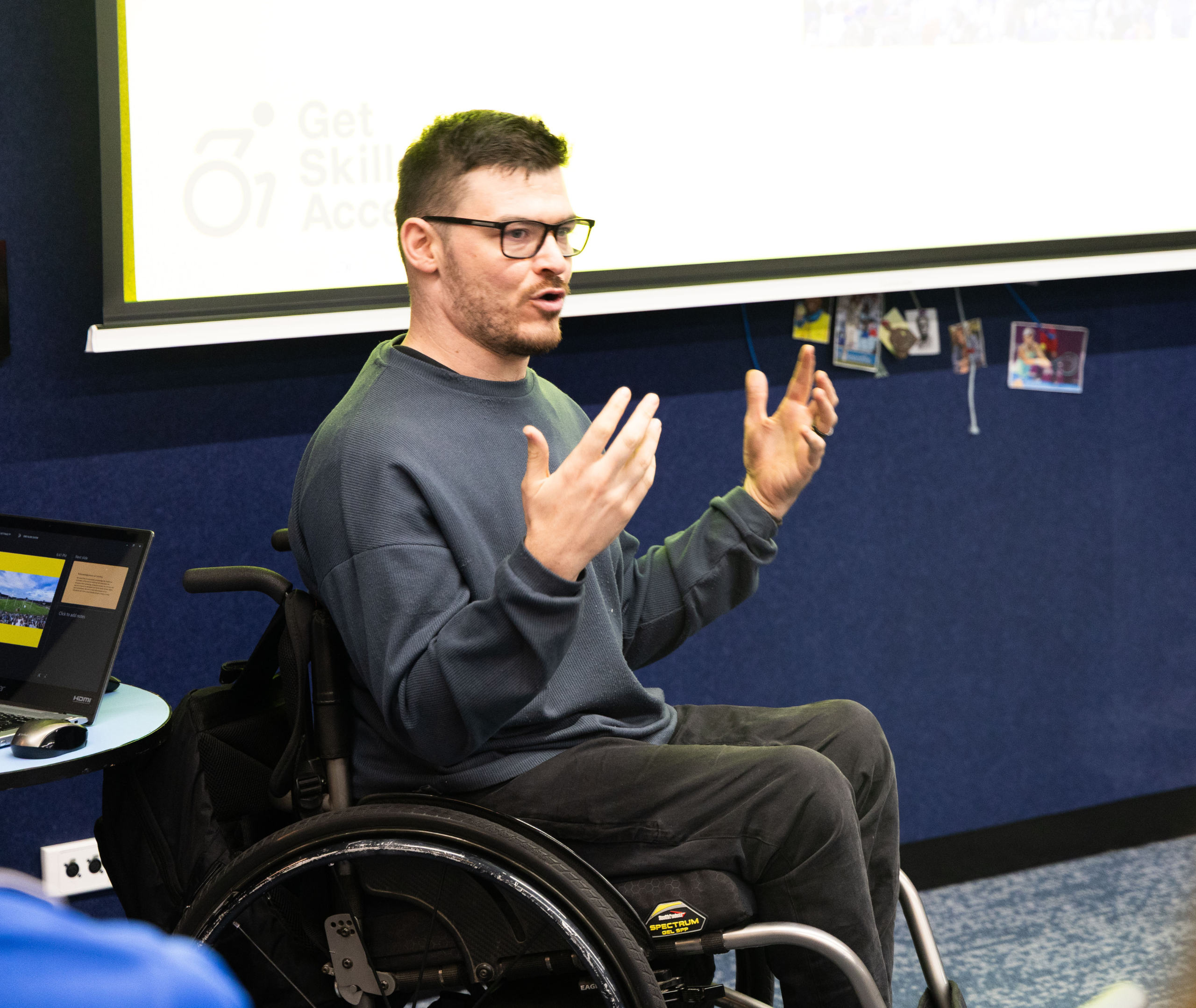 Sport4All Inclusion Coach, Oliver is on a wheelchair gesturing with his hands while speaking in front of a projected Get Skilled Access presentation slide.